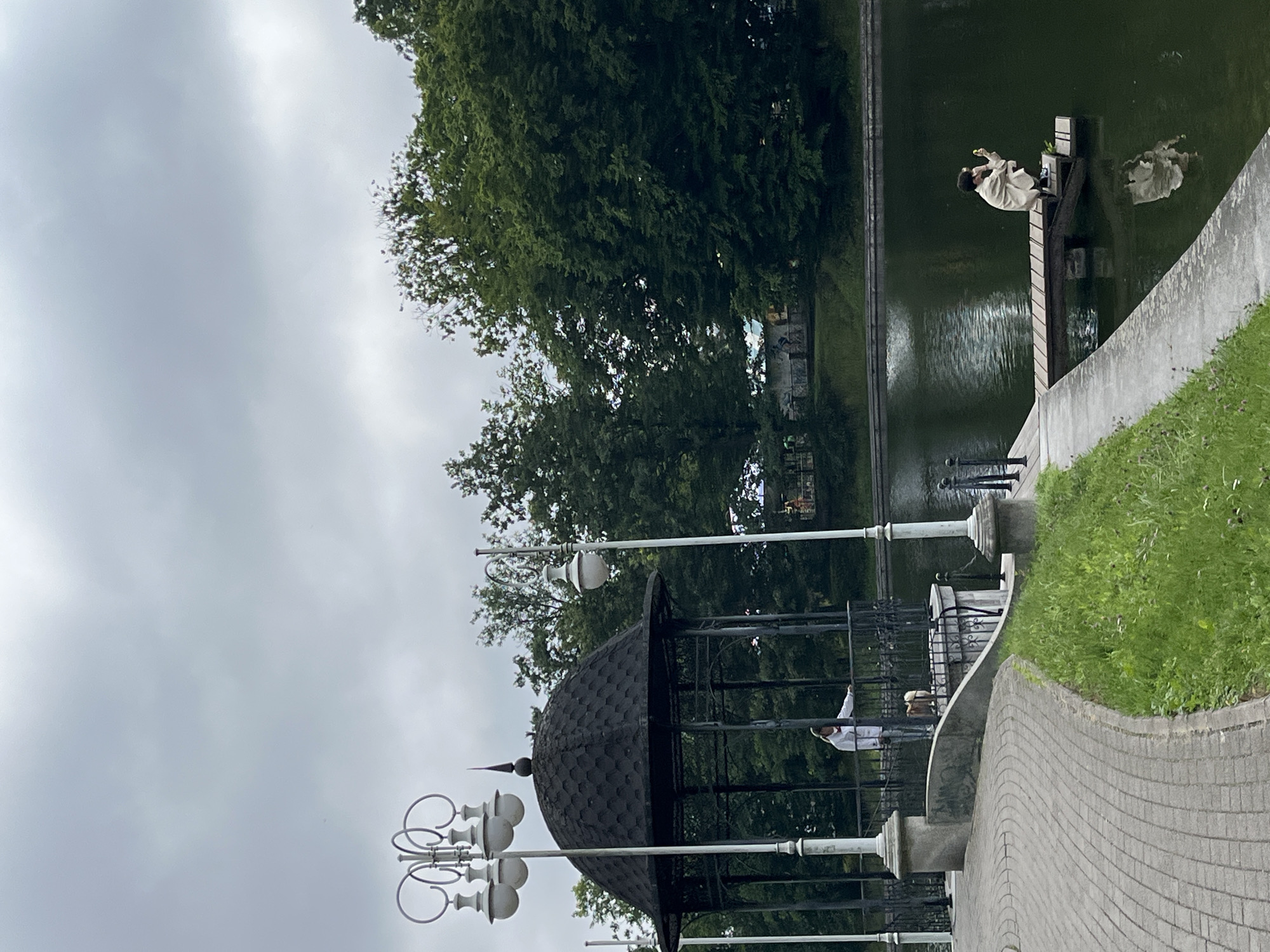 The image shows a park area with a pond. On the left, there is a gazebo with a dark, domed roof. A person in a light-colored outfit is standing inside the gazebo, leaning on the railing. A small dog is also present near the person. To the right of the gazebo, a person is sitting on a wooden pier extending into the pond, holding a phone. The pond is dark green. The background features lush green trees under an overcast sky. A paved pathway is visible in the foreground, curving towards the gazebo, with green grass bordering it. This scene is located in the city of Kaliningrad, Russia, specifically within the park surrounding the Upper and Lower Ponds.