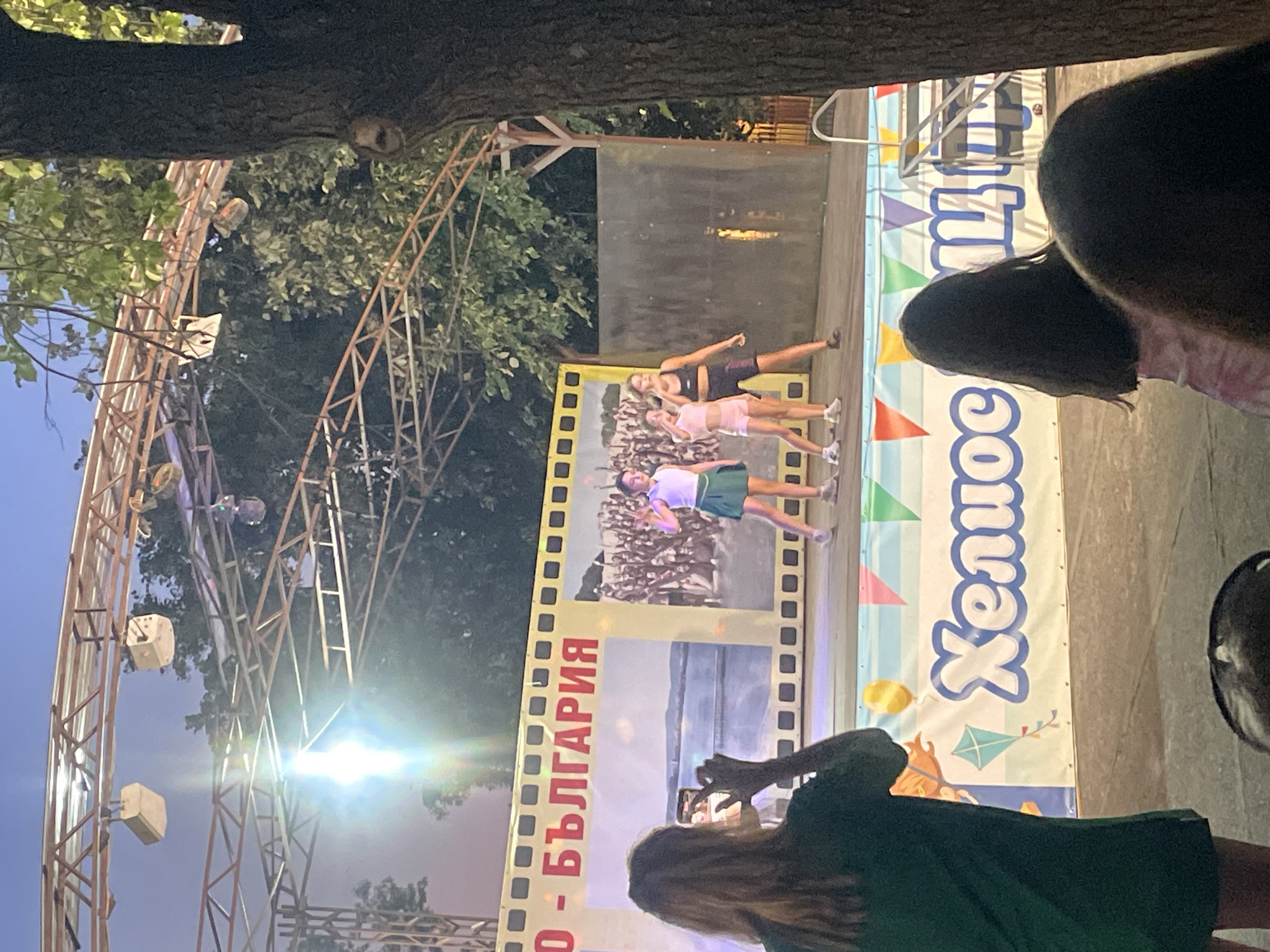 The image depicts an outdoor stage with a performance taking place at dusk. Three young women in athletic wear are posing on the stage, with one in the foreground and two behind her. A large backdrop behind them resembles a film strip and features images of people and a beach scene, with Bulgarian text that reads "- БЪЛГАРИЯ" (Bulgaria). To the right of the stage, a banner with colorful triangular patterns and text that appears to be in Cyrillic is visible.
In the foreground, a person with long dark hair, wearing a dark green dress, is holding up a smartphone to capture the performance. The scene is set under a large tree, with stage lighting illuminating the performers. The overall atmosphere suggests a lively event, possibly a festival or performance in a tourist area. The time of day is evening, as indicated by the dim natural light and the stage lighting. The presence of trees suggests a natural or park-like setting. The text on the backdrop and banner strongly indicates the location is in Bulgaria.