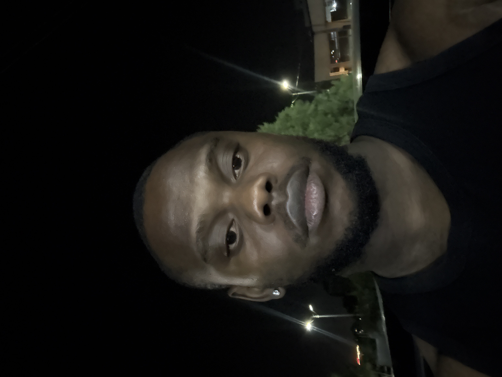A close-up, slightly off-center portrait of a dark-skinned man with a shaved head and a beard. He is wearing a black tank top. The background is predominantly dark, with streetlights visible in the lower part of the frame. A building and green foliage can be discerned in the background behind the subject. The picture was taken outdoors at night in Lugbe, Nigeria.