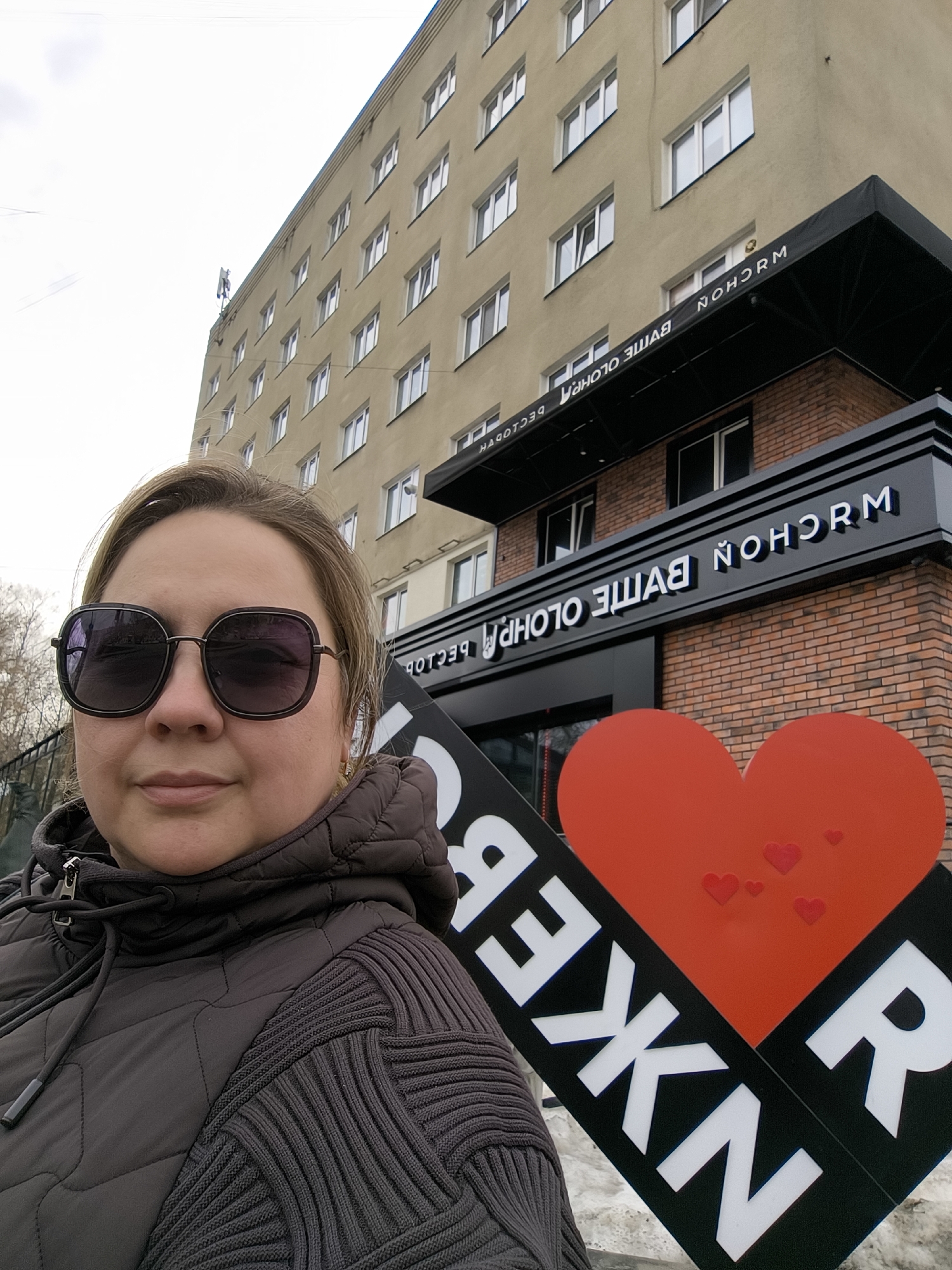 A woman wearing sunglasses and a brown quilted and knitted jacket is posing for a selfie in front of a building in Izhevsk, Russia. The building has a black awning with the name "NOHORM" displayed in white letters, both right-reading and mirrored. The word "PHOTO BATTLE" is displayed below the awning, also in white letters and mirrored. A large sign with a red heart and the backward letters "REKN" is held to the right. The building is tan and has several rows of windows. The sky is overcast. Bare trees are visible in the background.