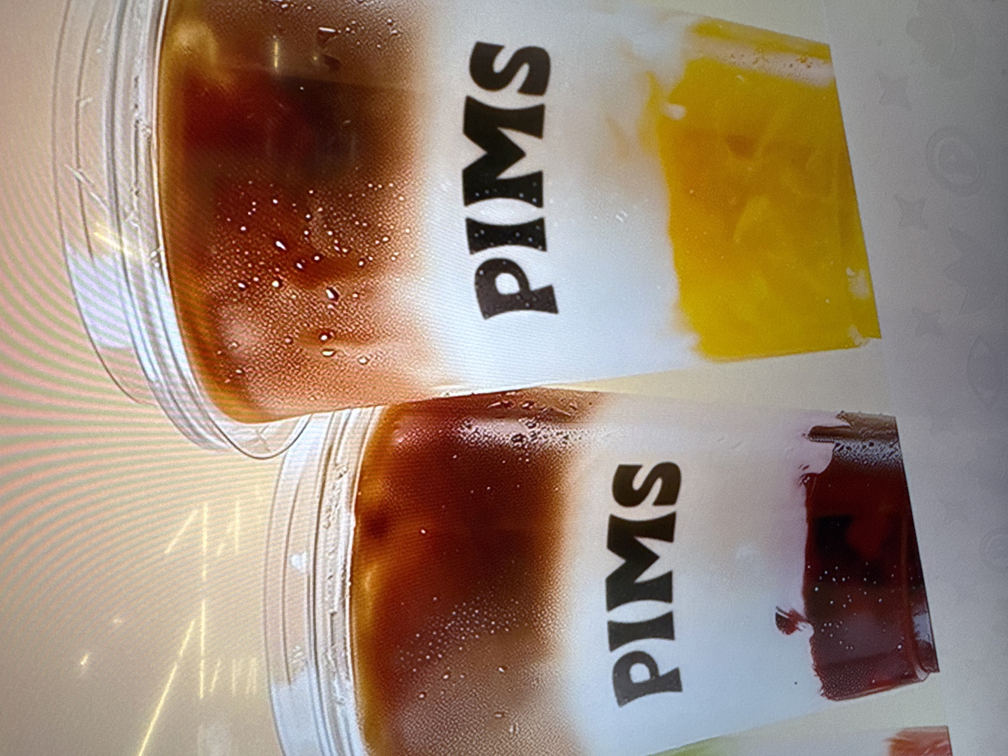 The image shows two plastic cups, each with a drink labeled "PIMS." The drinks appear layered, with a dark-colored liquid at the top, a white layer in the middle, and a colored layer at the bottom. One drink has a yellow bottom layer, while the other has a reddish-brown layer. The cups are sitting on a light-colored surface with reflections. There is no indication of the location in Moscow, Russia.