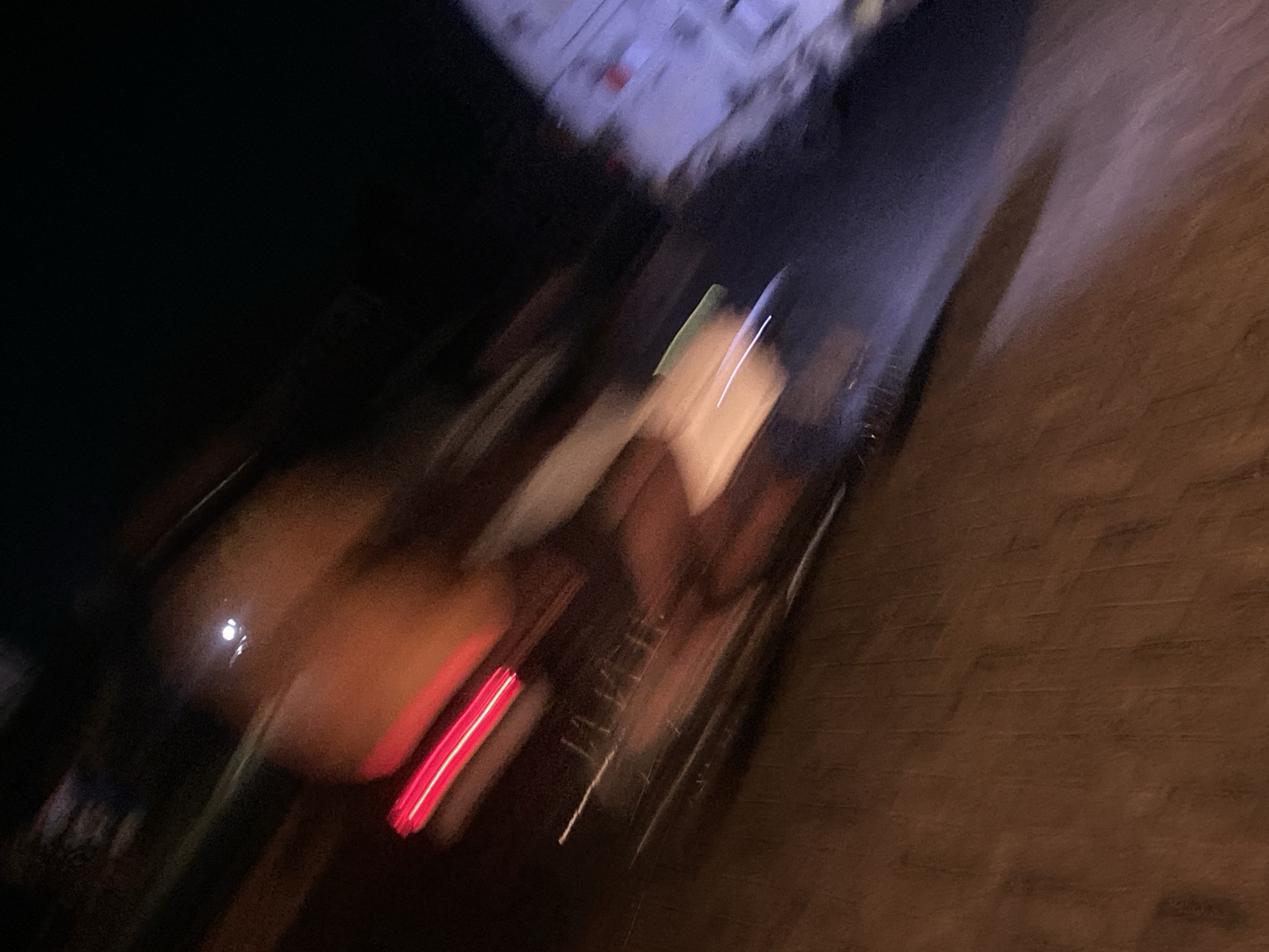 The image depicts a heavily motion-blurred outdoor scene at night in Ikorodu, Nigeria. The primary subject is a vehicle, likely a car or motorcycle, moving from left to right, indicated by the elongated streaking of light sources. A prominent, bright red horizontal streak, consistent with a vehicle's taillight, is visible on the left side. Above and to the right of this red light, a large, indistinct, orange-brown mass suggests the vehicle's body. Further up, a distinct small white circular light source is present.
The immediate foreground displays a textured, dark brown surface, likely pavement or ground, exhibiting vertical striations. To the right of the main vehicle, blurry white and green light streaks are observed against a darker background, possibly reflections on the ground or another part of the moving object. In the far right background, a large, indistinct, light-colored (off-white to light purple) object with some red marking is present, possibly a building or another stationary vehicle. The overall scene is characterized by low light conditions and significant motion blur, suggesting either high speed of the subject, camera movement, or a long exposure time.