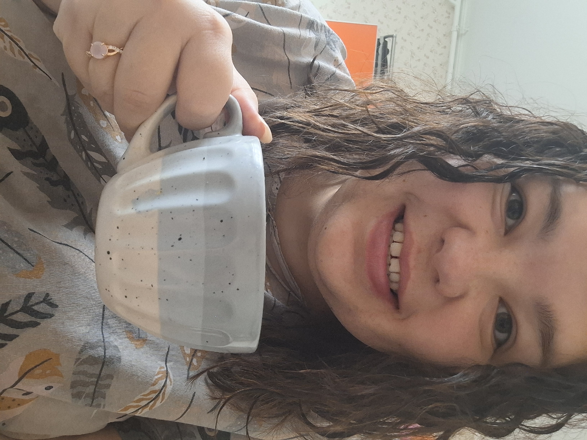 A close-up, rotated image shows a person, likely female, holding a two-toned ceramic mug while smiling. The perspective is from a low angle, with the subject appearing to be lying down or lounging, as her head is tilted and rotated counter-clockwise.
The subject has dark, curly or wavy hair that appears wet or styled. Her face is partially visible, showing fair skin, freckles on her nose and cheeks, and a wide smile revealing her upper teeth. Her right eye, dark in color, is clearly visible.
She holds the mug in her right hand, which also displays a gold-toned ring on the ring finger. The ring features an oval-shaped, light pink gemstone, possibly rose quartz, set between smaller clear stones on the band. The mug has a speckled white bottom and a speckled light grey top section.
The person is wearing or covered by fabric with a repeating pattern of stylized feathers or leaves in shades of grey, black, and mustard yellow on a light background. The background reveals a light-colored wall with faint texture, and in the upper right, a bright orange rectangular object and a dark vertical shape, suggesting an indoor setting like a room. No visible text is present. The smile conveys a sense of happiness or relaxation in this indoor scene in Markovskiy, Russia.