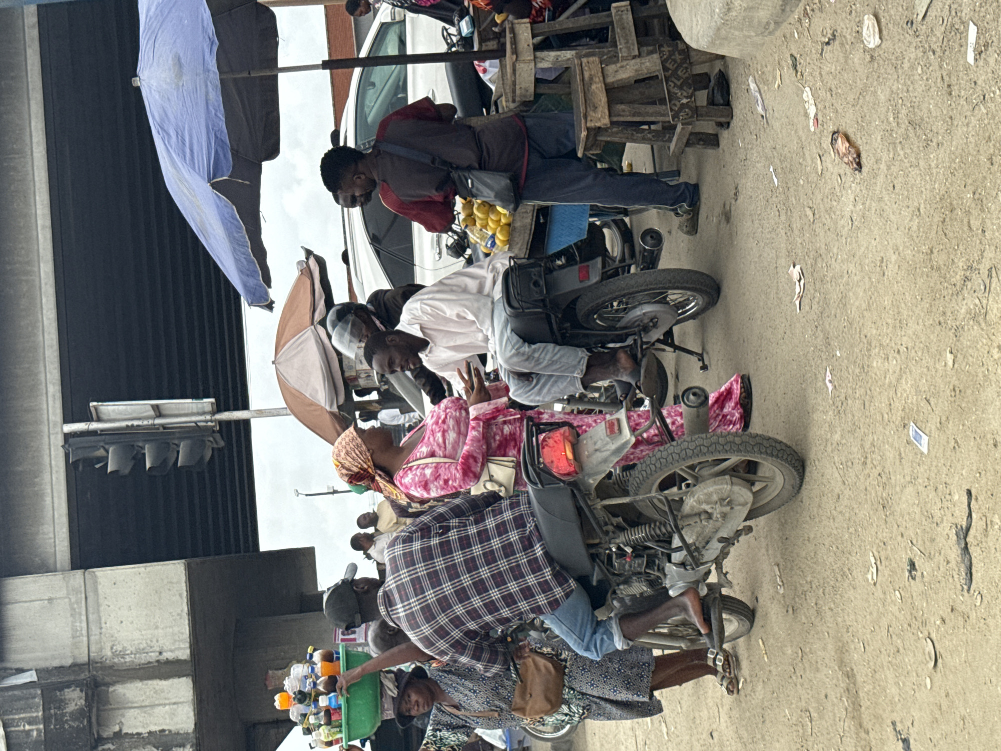 The image captures a bustling street scene in Lekki, Nigeria, beneath an elevated highway. In the foreground, a man in a plaid shirt is seated on a motorcycle, facing right. To his left, a woman wearing a patterned dress and a headscarf carries a tray of drinks on her head. She is interacting with another person whose back is to the camera. To the right of the motorcycle, a man in a light-colored shirt is also seated on a motorcycle, facing left, and appears to be engaged in conversation with a man standing beside him, who is wearing a dark jacket and holding a bag. A stall selling fruits is visible in the background, with oranges displayed on a table. Two large umbrellas, one blue and one a muted orange and brown, provide shade. The ground is dirt and littered with debris. The overall atmosphere suggests a busy marketplace or a roadside stop.