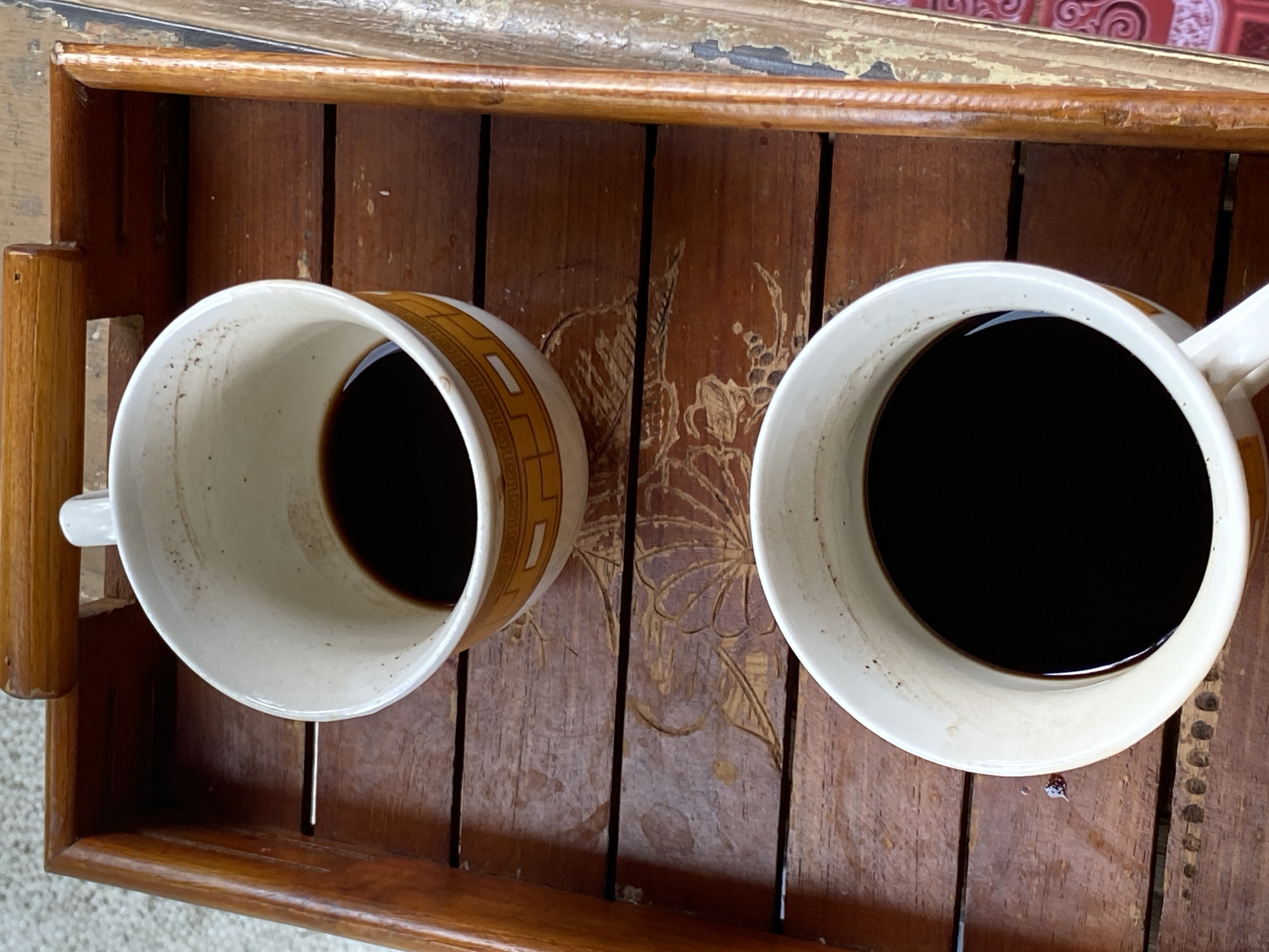 Two white ceramic mugs filled with dark liquid, presumably coffee, are placed on a rustic wooden tray. The mugs have a patterned design around the rim, with the top mug showing a yellow and white geometric pattern. The wooden tray has a weathered appearance with visible grain and some decorative carvings. The tray is placed on a textured surface, possibly carpet or a mat, with a blurry background. The scene is indoors, likely in a domestic setting, given the presence of the tray and mugs. The image does not provide any specific information about the location within Lambaro Kueh, Indonesia, beyond the implication that such a scene could be found there. There are no people, actions, or interactions depicted.