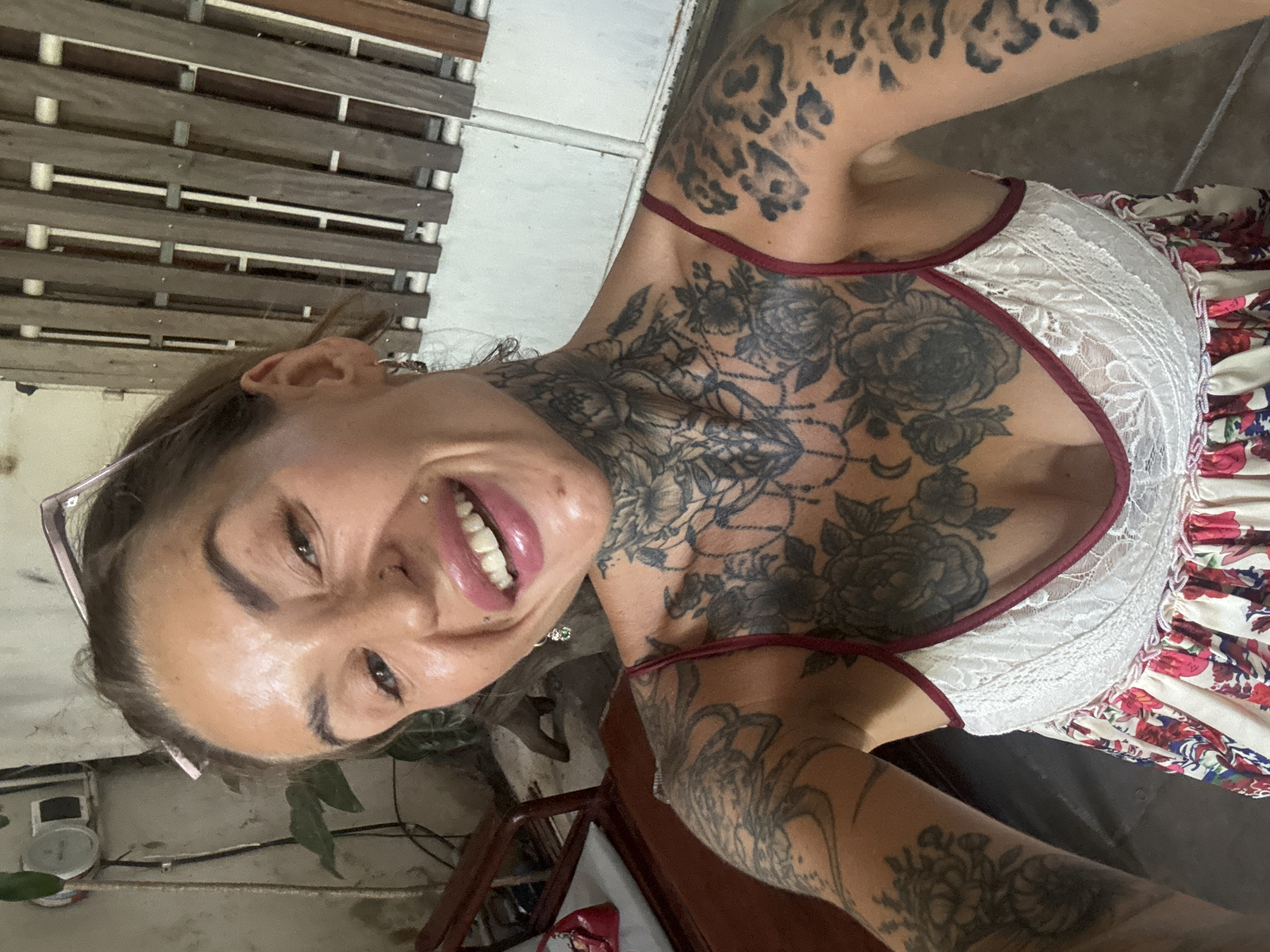 A woman with extensive tattoos on her neck, chest, and arms takes a selfie outdoors. She is wearing a white and floral print dress with lace detailing. She has her hair pulled back and is wearing glasses on top of her head. She is smiling, showing her teeth, and has piercings on her face and lips. The background appears to be a rustic outdoor setting, possibly a patio or garden area, with a wooden fence and some greenery. There is a chair with a red bag on it to her left. The location is identified as Bac Nha Trang Ward, Vietnam.