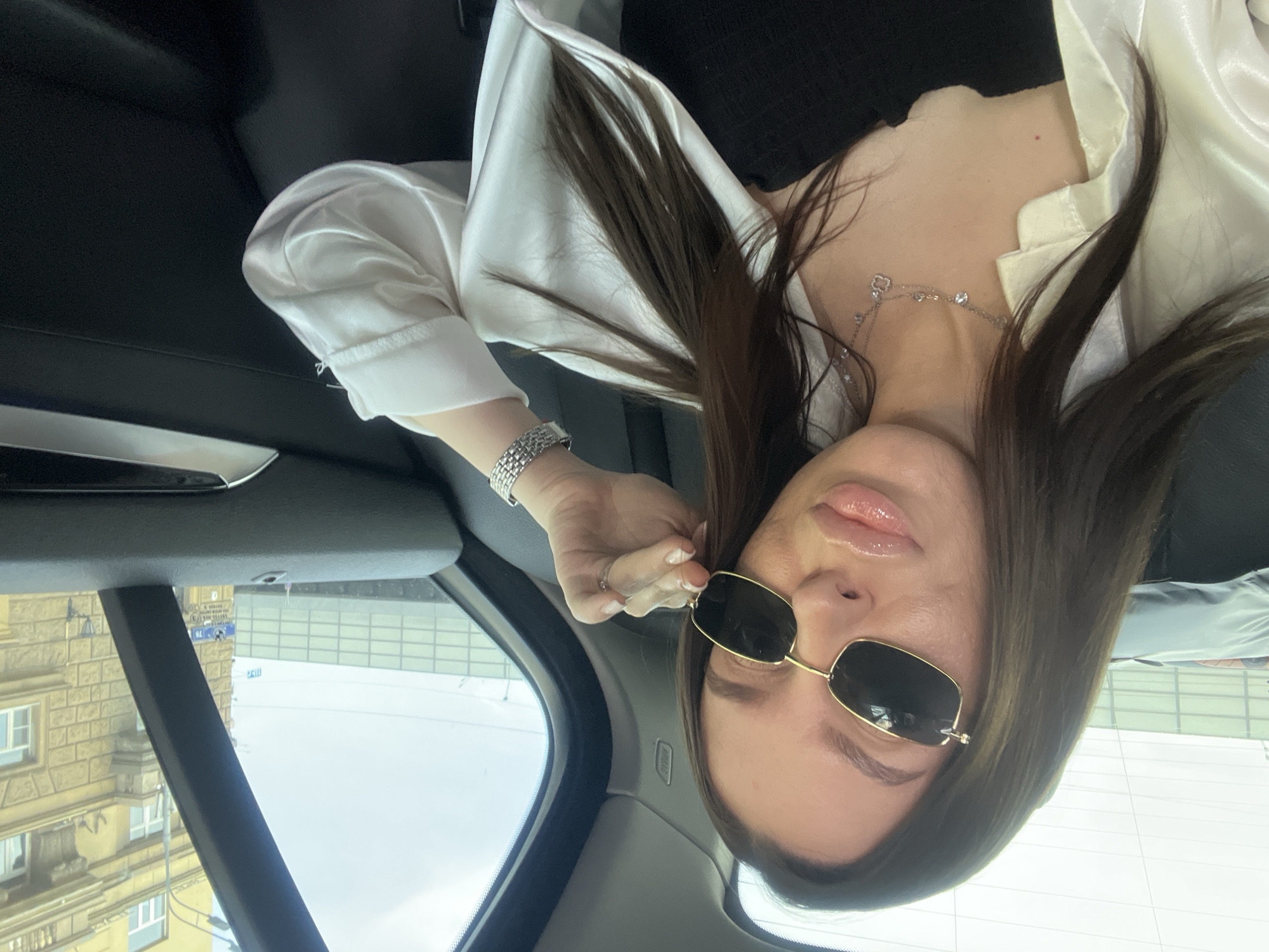 A woman wearing sunglasses and a white blouse is sitting in a car in Moscow, Russia. She is facing the camera. The building outside the car window contains a blue street sign with the address of Дмитровский пер. дом 7/6, стр. 1а.