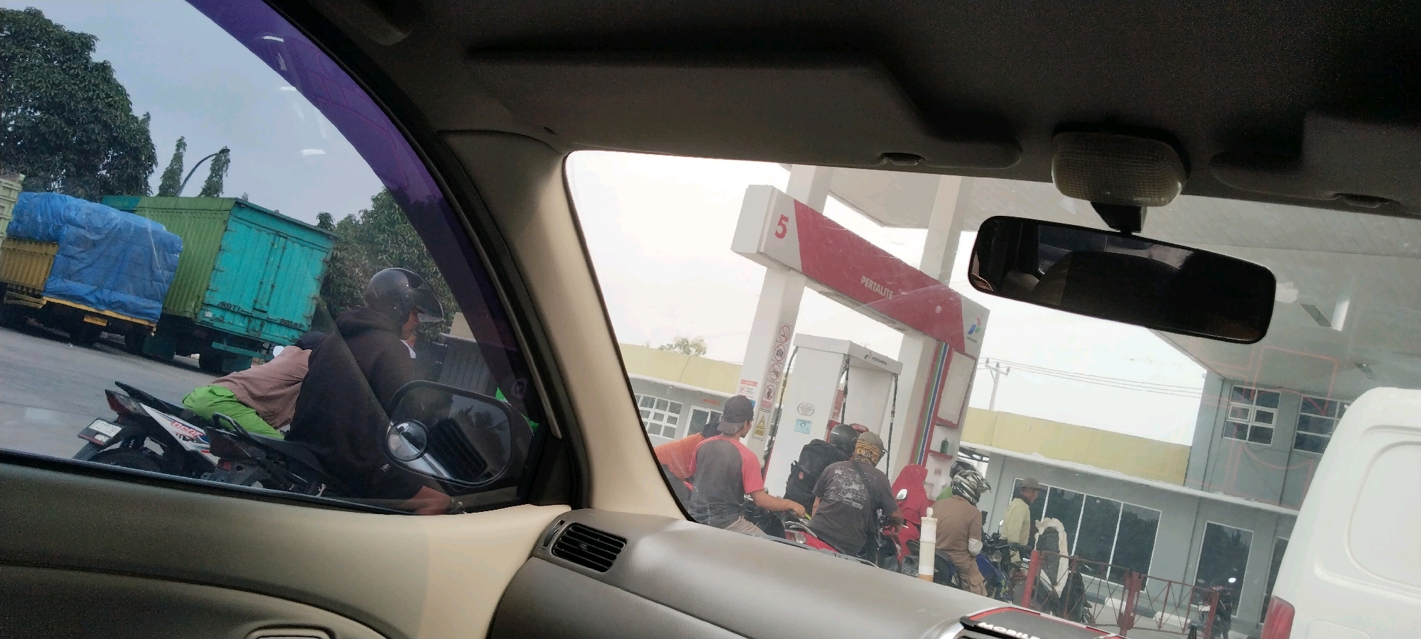 A wide shot captures a scene from inside a vehicle, looking out at a Pertamina gas station in Haduyang, Indonesia. The sky is overcast, and trees line the background. In the foreground, through the car's window, a green cargo truck covered with a blue tarp is parked on the left. Several people on motorcycles are gathered around the gas pumps. One person is wearing a black helmet and a brown hooded jacket, leaning forward on their motorcycle. Another individual, wearing a brown top and green pants, is also on a motorcycle. Further into the scene, more individuals are on motorcycles, some are wearing helmets and casual clothing. The gas station structure is prominent, with a red and white sign displaying "PERTALITE." Buildings with windows are visible behind the gas station. The overall atmosphere suggests a busy, everyday scene at a local service station.