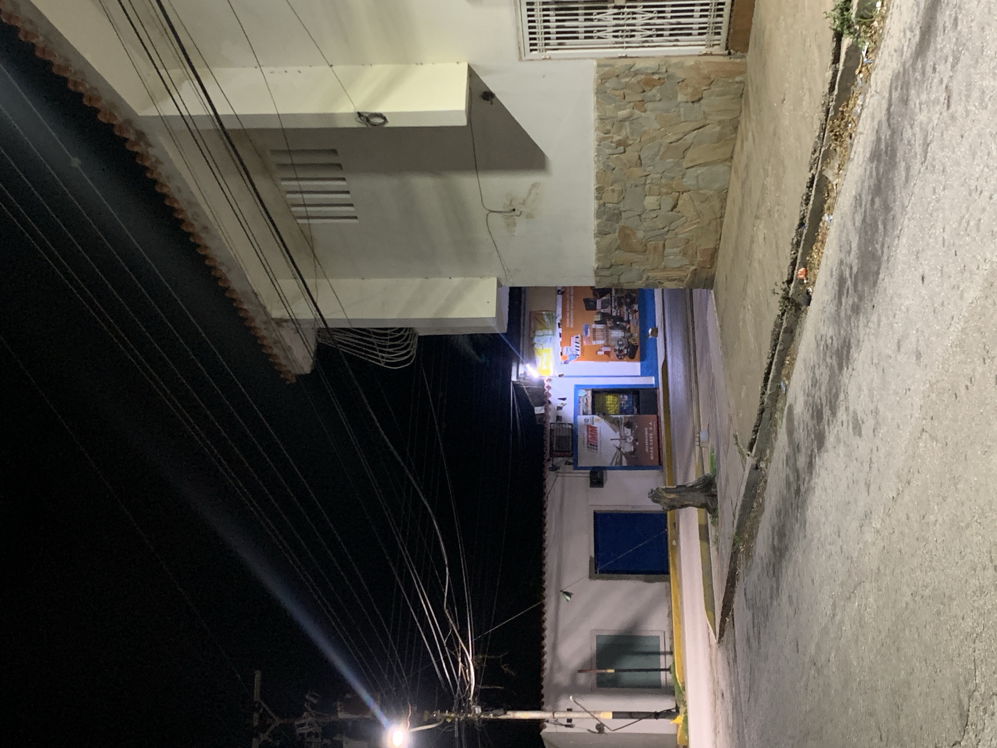 The image captures a nighttime street scene in Santa Ana, Venezuela. A streetlight illuminates a section of the road and the facade of a building. The building on the right has a stone base and a white upper story with a patterned window. To the left, another building features a blue door and a green-tinted window. Wires crisscross the dark sky above. A lone tree stump stands near the curb. Debris is scattered along the edge of the road and sidewalk. The overall impression is one of a quiet, sparsely populated urban environment at night.