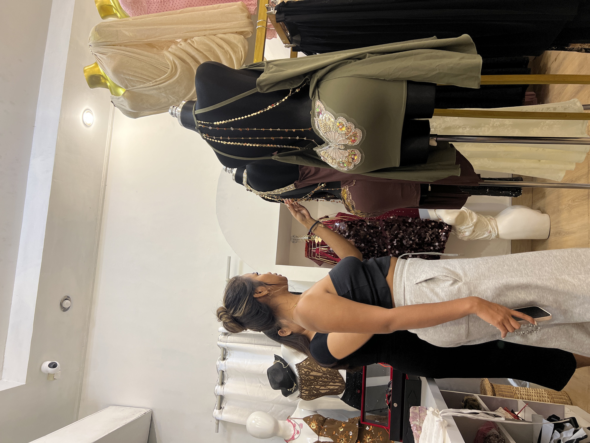 The photo shows a woman in a clothing store in Kuta, Indonesia. She is wearing a black tank top and light grey sweatpants. The woman is holding a cell phone in her right hand and looking at a clothing rack. The clothing rack contains a variety of dresses and outfits displayed on mannequins. The rack is placed against a white wall. Other objects in the store include additional mannequins and clothing racks, baskets, a security camera, and various garments for sale.