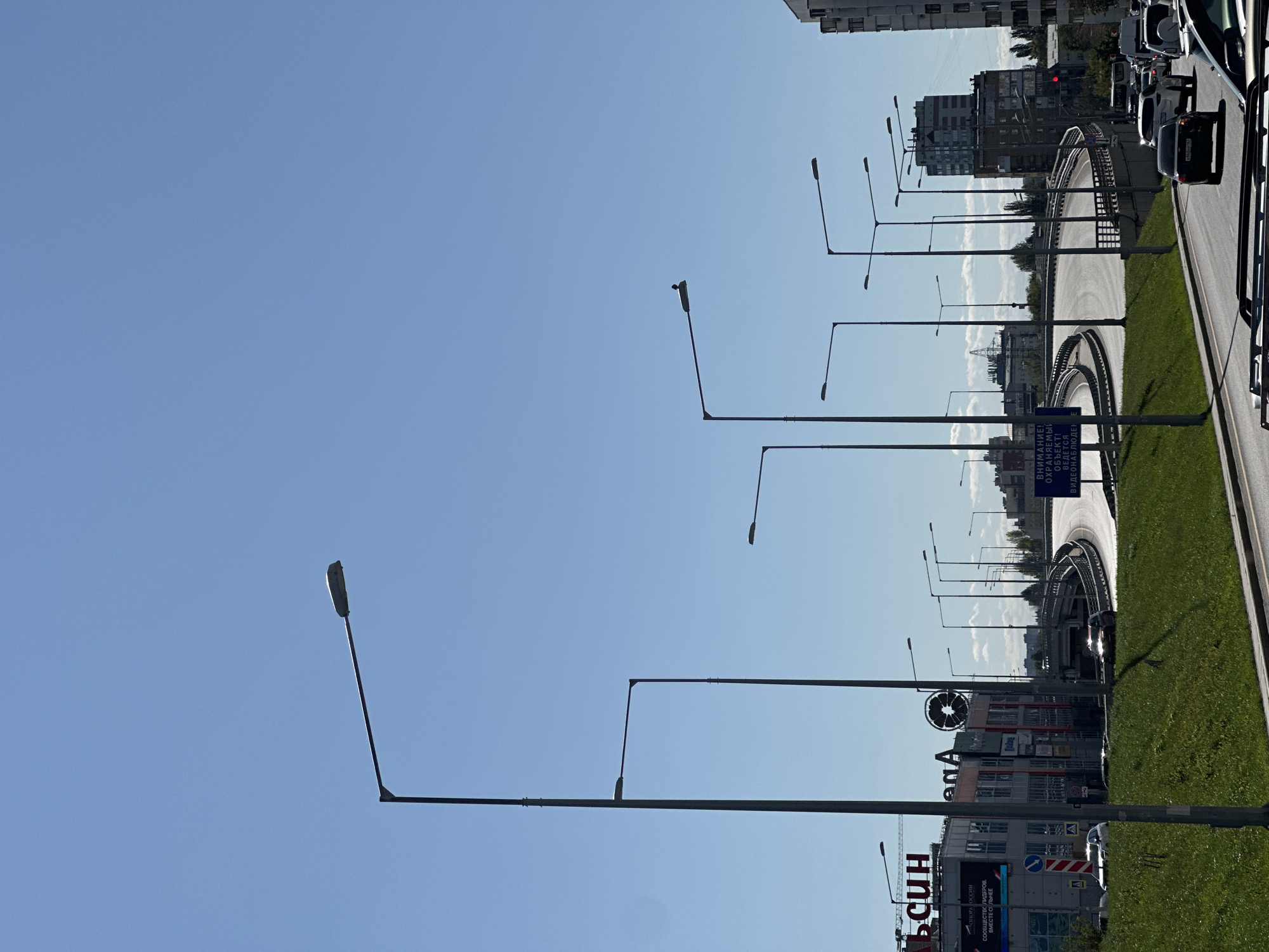 A wide shot captures a clear blue sky over a multi-lane highway overpass in an urban environment. Numerous streetlights line the roadway, casting shadows on the green grass median. Buildings, including a multi-story structure with advertisements, are visible on the left side of the frame. Cars are seen driving on the highway and on the lower road. A bird is perched on one of the streetlights. The scene appears to be a daytime shot.