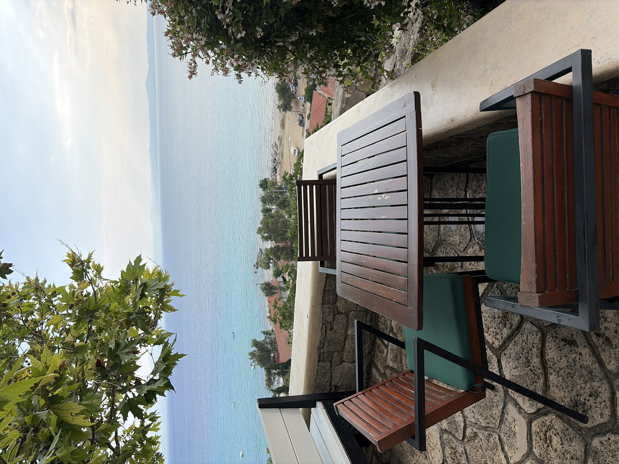 This is a full, eye-level shot of an outdoor seating area overlooking the sea in Kassandra Municipal Unit, Greece. The image features a dark wood table with a slatted surface, supported by black metal legs. Two identical wooden chairs with dark metal frames and green cushions are positioned at the table. A third wooden chair with a green cushion is visible behind the table, facing the sea. To the left, a white and wooden bench is partially visible.
The seating area is set on a stone-tiled patio. A low, rough-hewn stone wall separates the patio from a concrete ledge. Beyond the ledge, the turquoise sea stretches to the horizon, meeting a hazy sky. In the distance, a coastline with scattered red-roofed buildings and green trees is visible. Several small boats are dotted on the water. Lush green foliage from a large tree hangs over the top left of the frame, and a flowering shrub with small pink and white blossoms is prominent on the right. The overall impression is a serene and inviting outdoor dining or relaxation space with a beautiful ocean view.