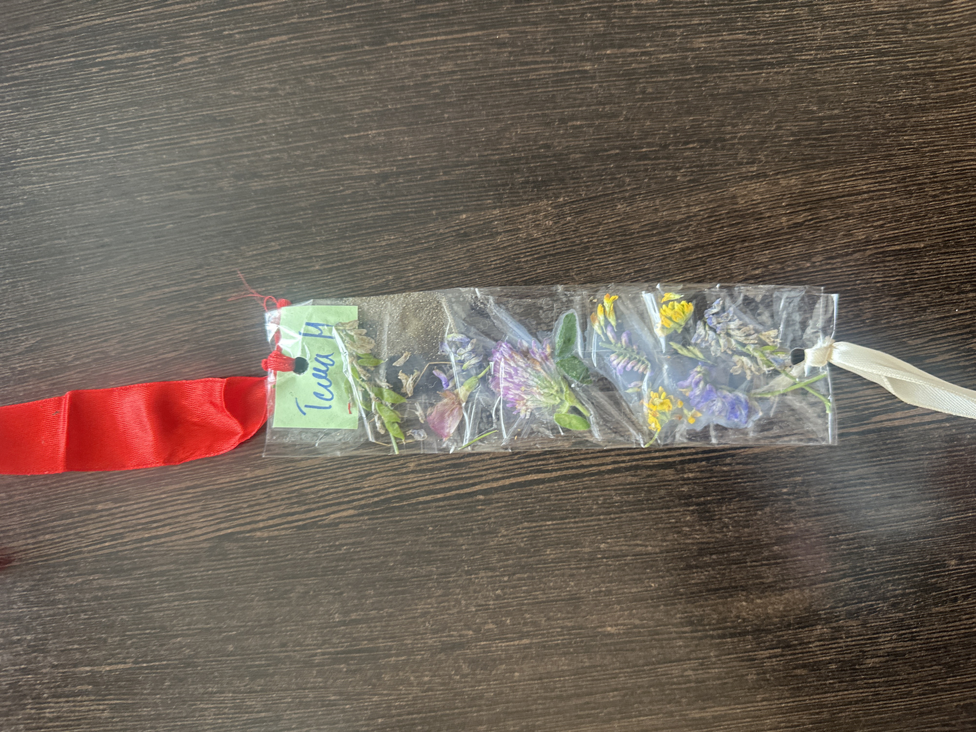 The image shows a bookmark made of pressed flowers encapsulated in plastic. A red ribbon is attached to the top of the bookmark, and a light-colored ribbon is attached to the bottom. A small green label with blue writing, possibly a name or title, is affixed to the top of the bookmark. The bookmark is laid flat on a dark wooden surface. The location in the city of Novoaltaysk, Russia is unknown as there are no identifying features present in the image.