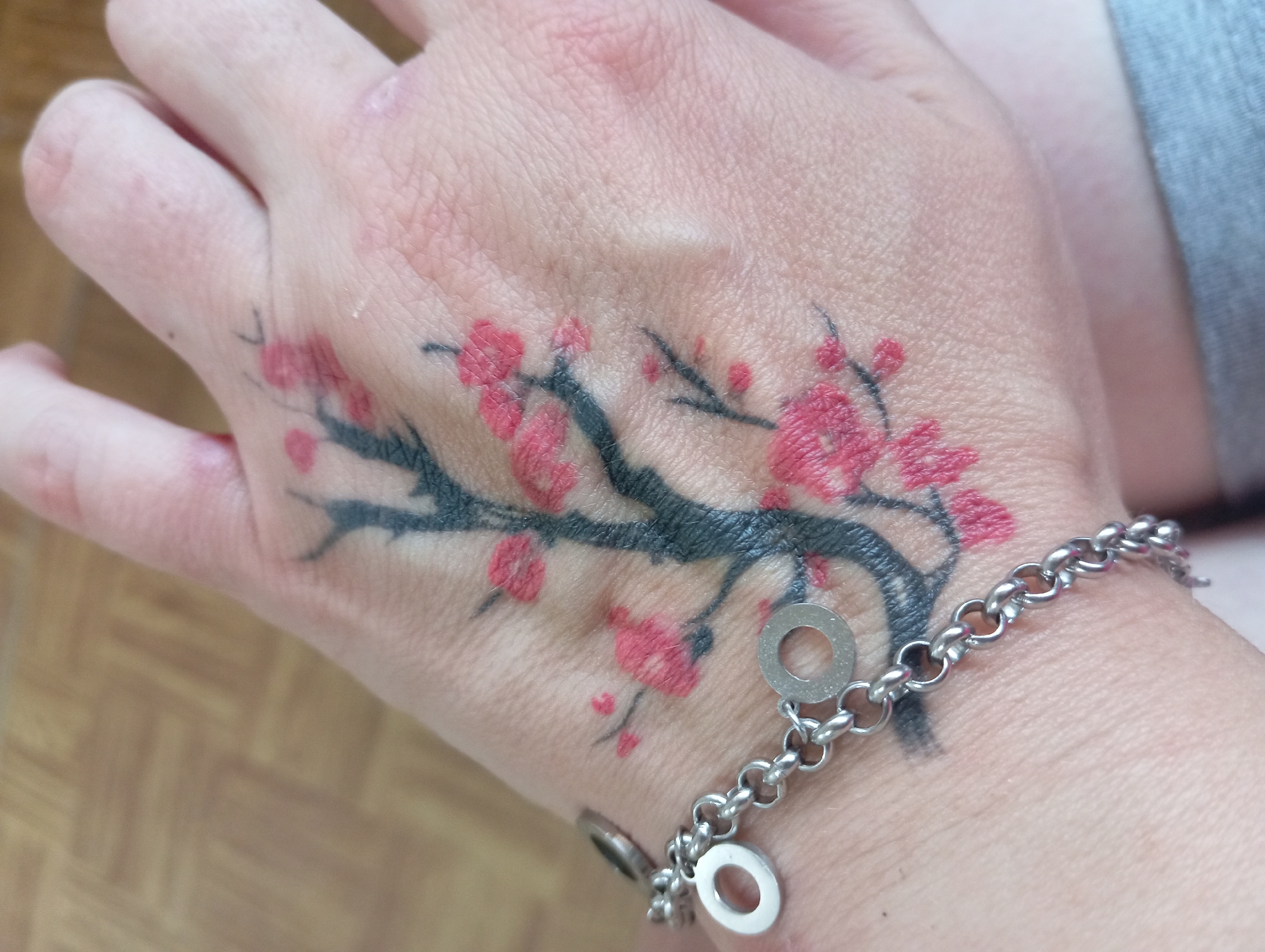 The image shows a close-up of a person's left hand, which has a tattoo of a cherry blossom branch with pink flowers and black branches. A silver bracelet with circular charms is worn on the wrist. The background is a blurred wooden floor. The location is not specified in the image.