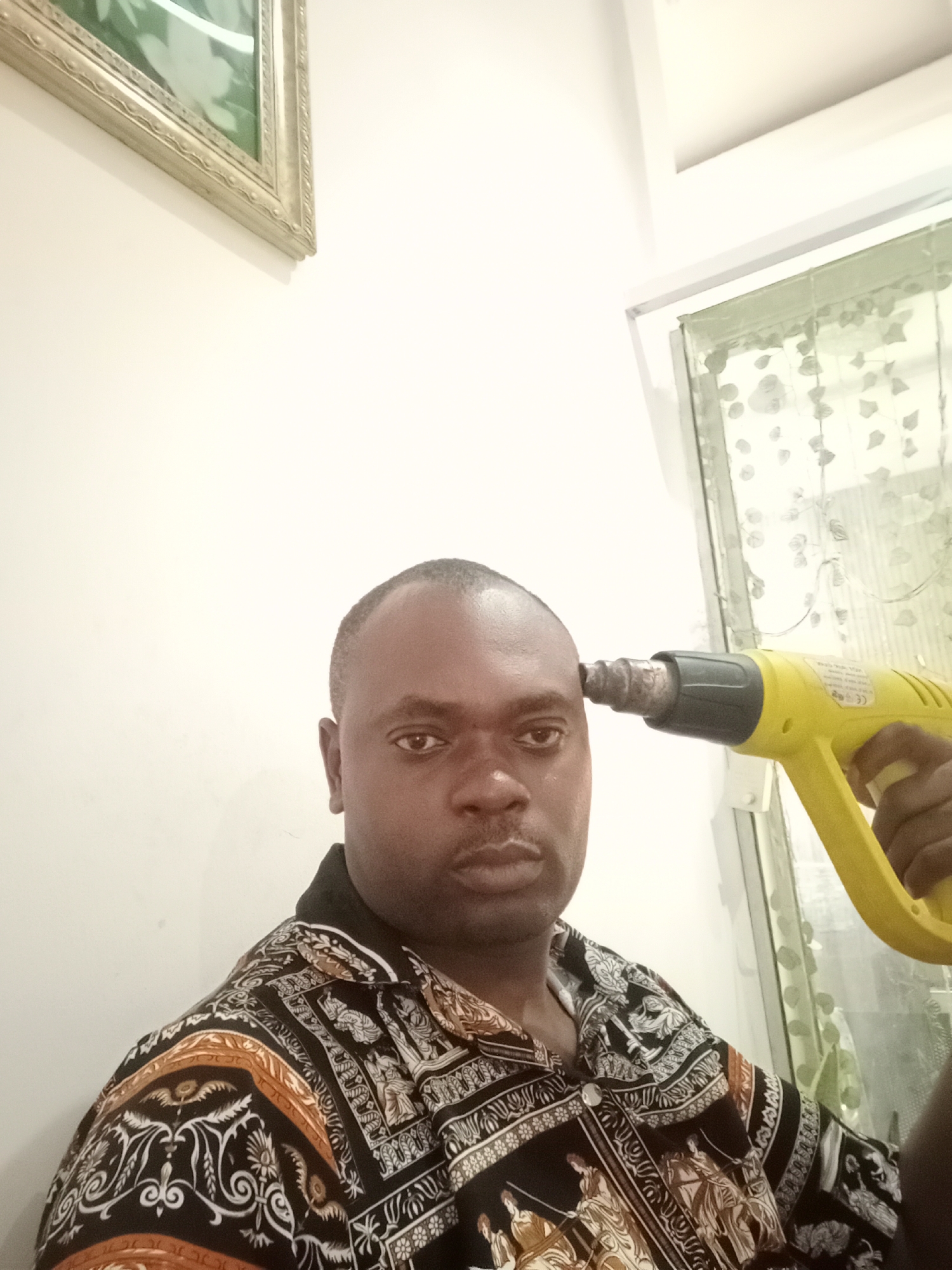 A man is pictured from the chest up in a room in Gwarinpa, Nigeria. He is Black, has a shaved head, and is wearing a patterned shirt. A yellow heat gun is being held next to his head, pointing toward his temple. Behind him is a window with a curtain. A picture hangs on the wall above and to the left.
