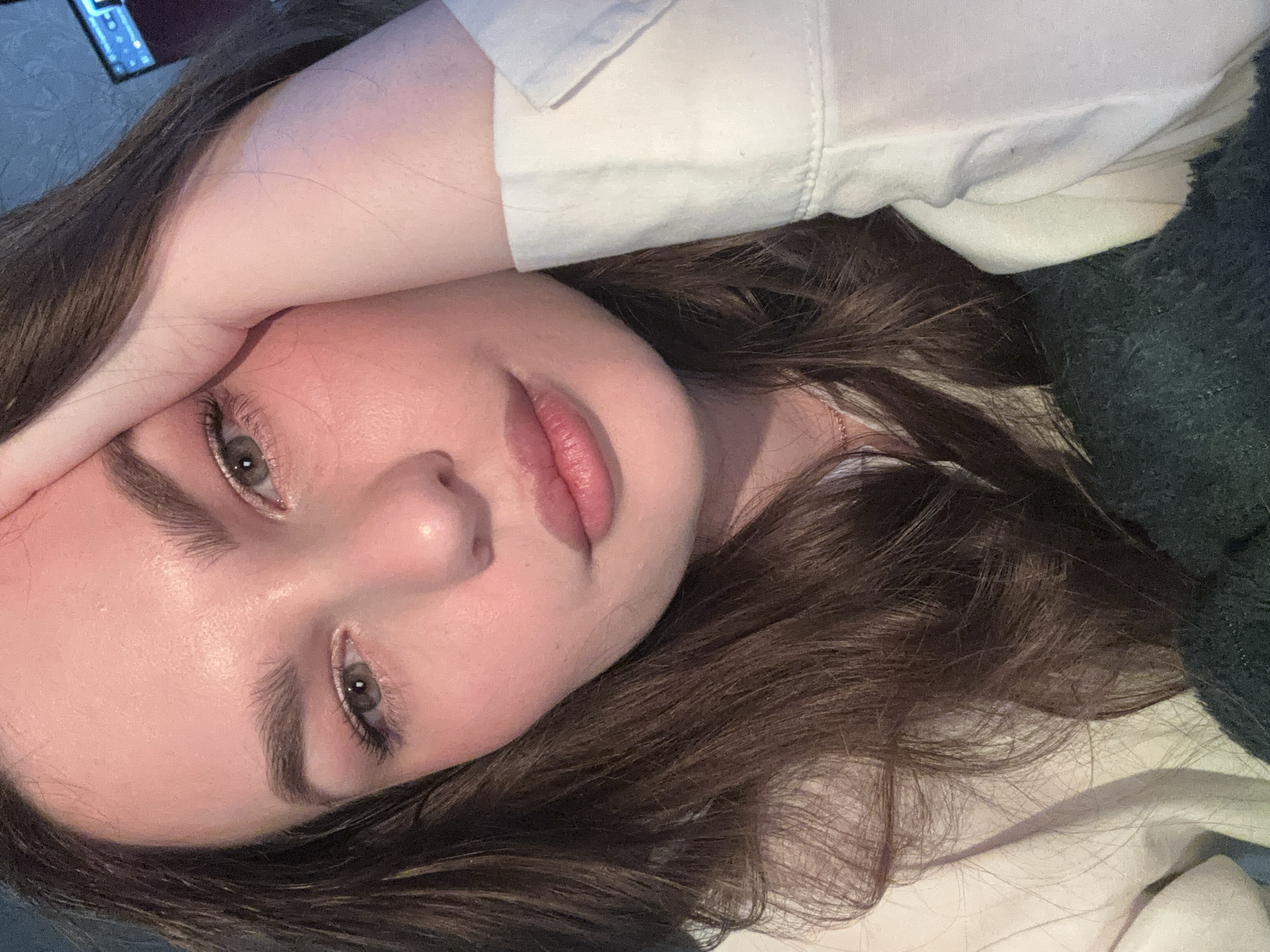 A close-up, slightly angled shot features a young woman with fair skin, brown hair, and green eyes. She is wearing a white shirt or blouse. Her left hand is raised and partially obscures her forehead. The background is indistinct, showing dark green and gray textures, suggesting bedding or fabric. The lighting appears to be soft and diffused. There is no information in the image to suggest the location is Chaikovsky, Russia, or any specific city.
