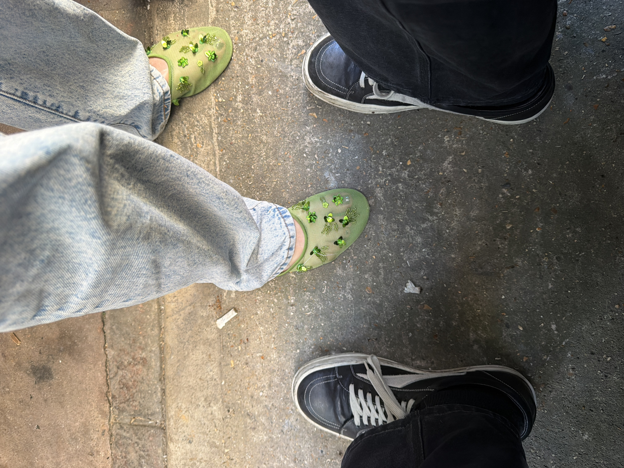 The image depicts a ground-level view of two pairs of feet standing on a paved surface. The pair on the left wears light-wash denim jeans and green mesh shoes adorned with green floral and sequin embellishments. The pair on the right wears black denim pants and black and white low-top sneakers. The paved surface appears to be a sidewalk or walkway. There is no indication of the city of London, United Kingdom. The scene contains no indication of actions or interactions.