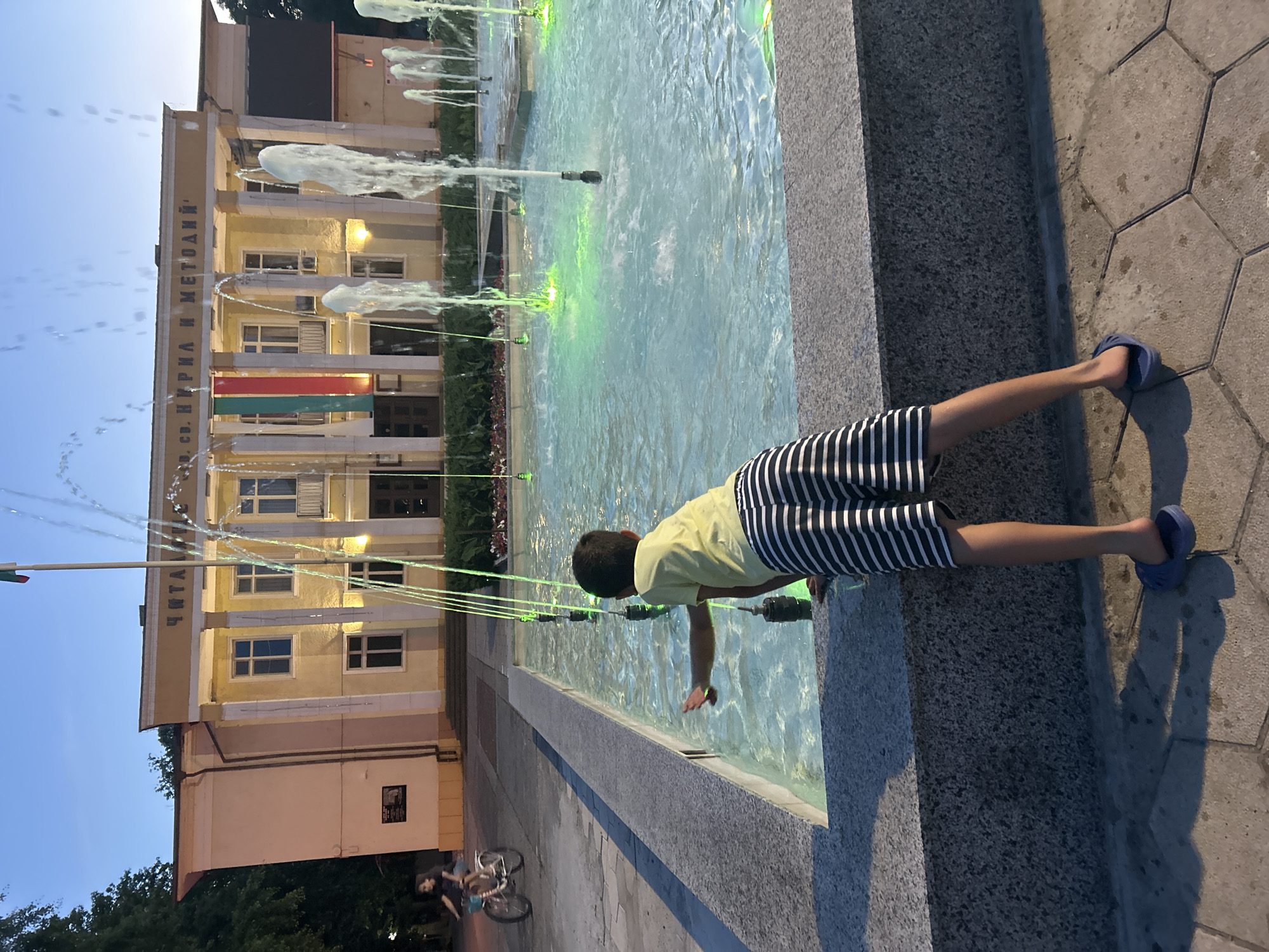 In Parvomay, Bulgaria, a boy in a yellow t-shirt and striped shorts leans over the edge of a lit fountain, reaching out to touch the water. The fountain features numerous jets of water, some illuminated with green light. In the background, a yellow building with columns and a sign that reads "ЧИТАЛИЩЕ СВ. КИРИЛ И МЕТОДИЙ" stands prominently. A flagpole with the Bulgarian flag is visible above the building. To the left, a young girl rides a bicycle on a paved area. The scene appears to be set during dusk, with a clear sky and trees visible. The ground around the fountain is paved with hexagonal tiles.