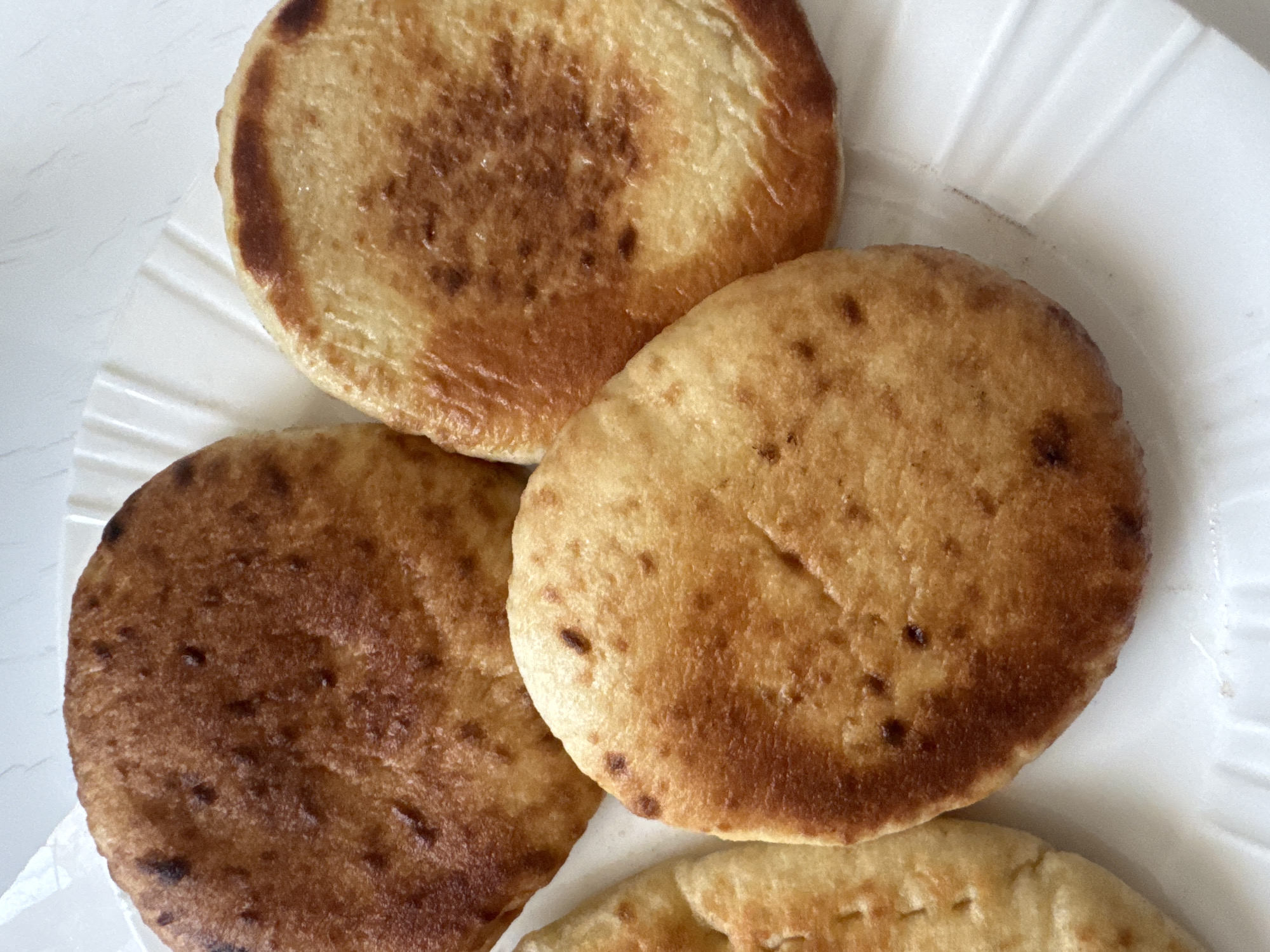 The image shows a close-up of four fried bread items on a white, ridged plate. The bread items are circular or oval in shape and have a golden-brown color with some darker, slightly charred spots. The texture appears slightly puffed and uneven. The background is a plain white surface. The scene is indoors and lit by overhead lighting. The bread items are arranged in a cluster on the plate, with some overlapping. There are no people or other objects visible in the image. There is no information about the specific location in Agios Tychona Community, Cyprus, as the image only depicts food.