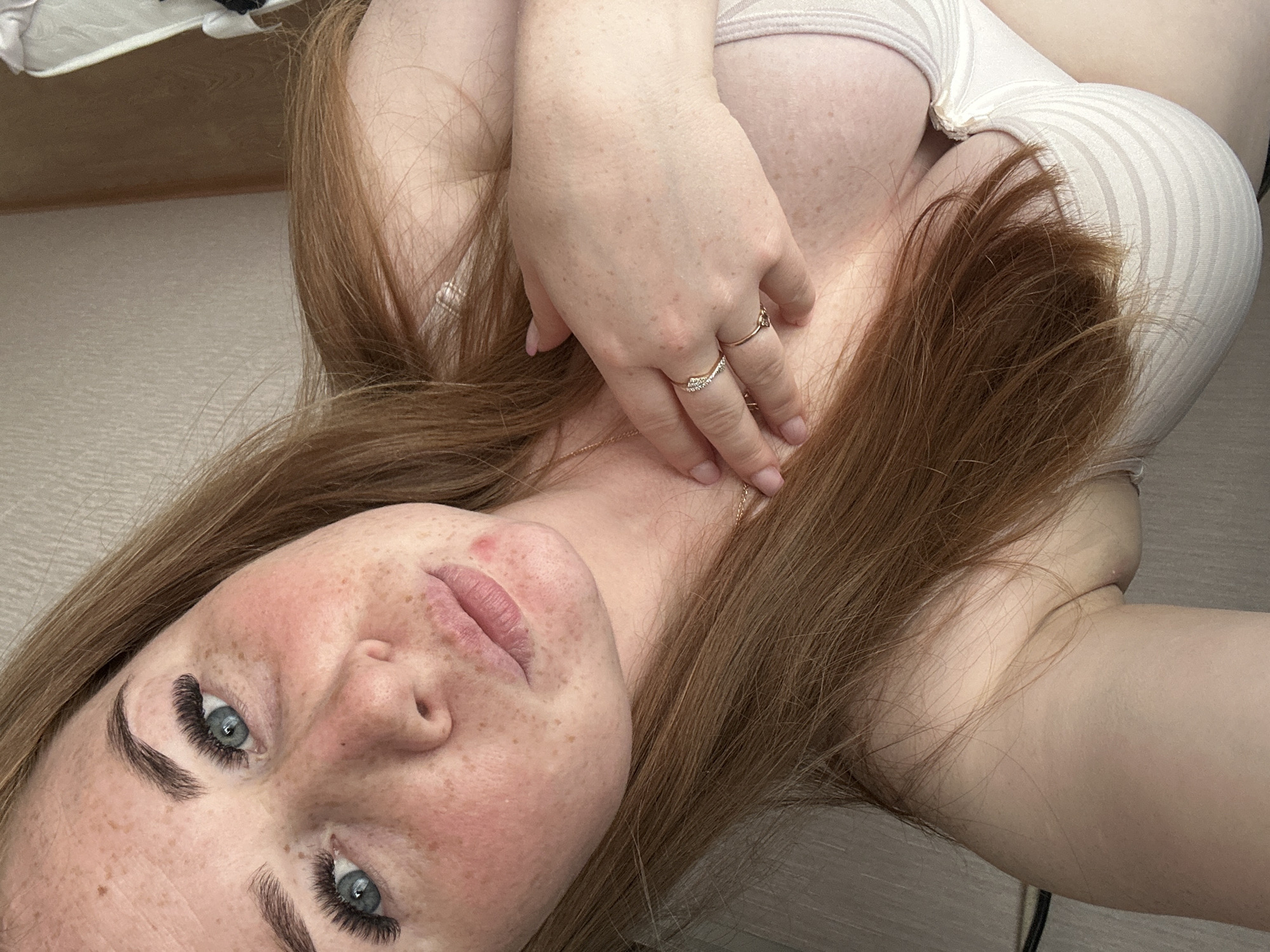 A woman with freckles and long auburn hair is lying down, looking up at the camera. She is wearing a light-colored top and rings on her fingers. Her hand is placed near her neck. The background appears to be a floor and potentially a bed or another surface. There is no visible information to indicate the location is in Chaikovsky, Russia.