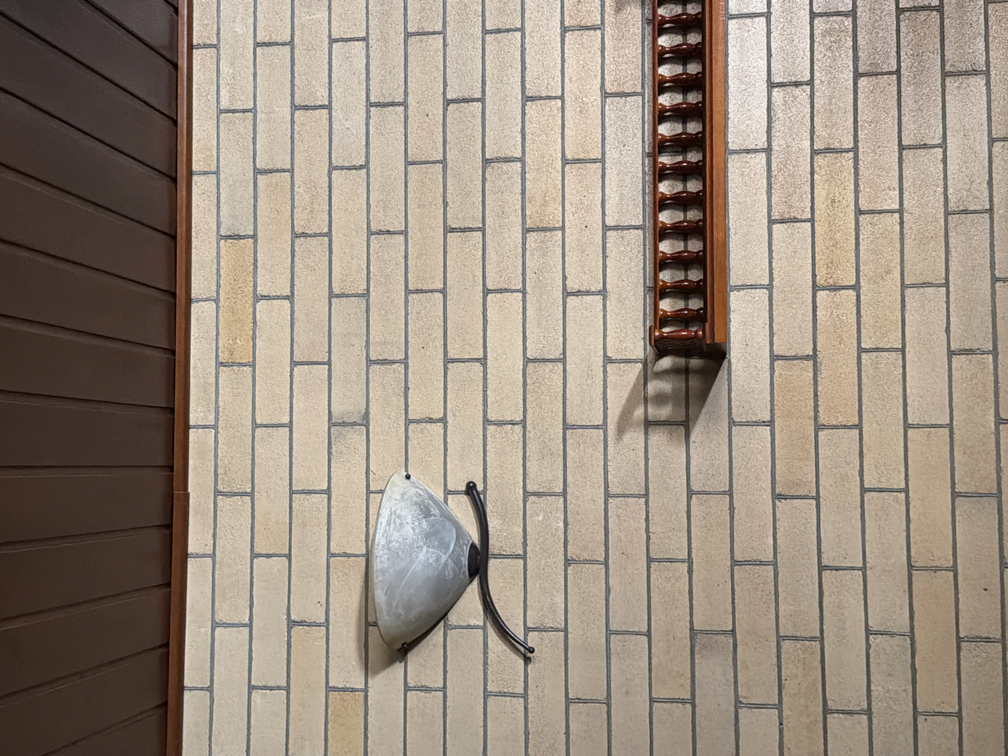 The image shows a brick wall in Qarağandı city administration, Kazakhstan. The wall is made of light-colored bricks with dark grout. There is a sconce mounted on the wall, and a wooden shelf with decorative spindles is positioned below it. The ceiling, visible at the top of the image, is made of dark wooden planks. The overall scene appears to be indoors.
