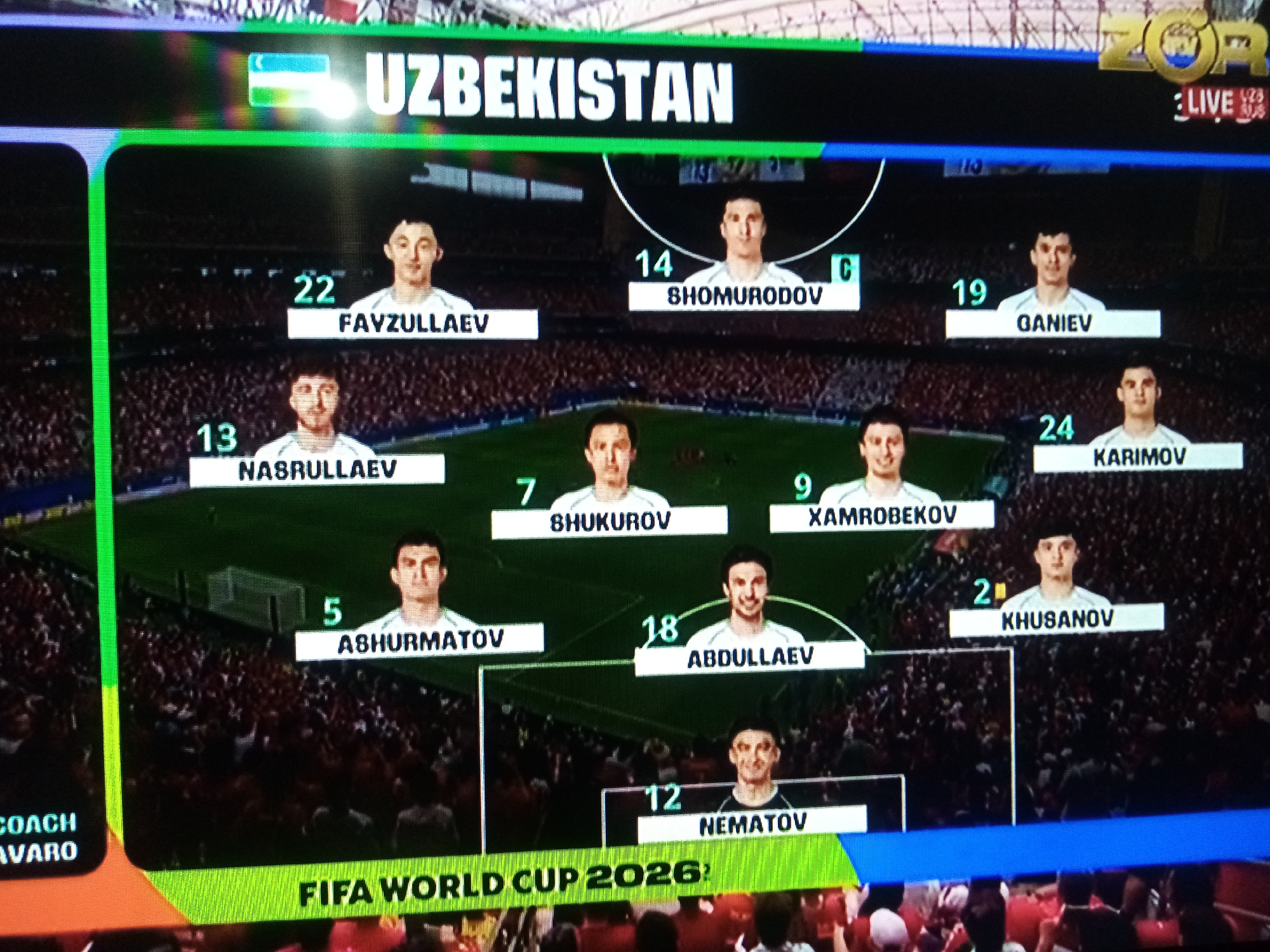 This is a screenshot of a soccer team's lineup display, likely from a video game or broadcast. The screen shows the "UZBEKISTAN" national team, with individual player portraits and their jersey numbers and names displayed. The players are arranged in a formation, suggesting a match is either about to begin or is in progress. A "LIVE" indicator is visible in the top right corner, along with a logo that reads "ZOR TV". The "FIFA WORLD CUP 2026" is also prominently displayed at the bottom, indicating the tournament context. The scene is set in a stadium, with the audience visible in the background, though out of focus. The overall atmosphere appears to be professional and related to a sporting event. Text includes player names such as "FAVZULLAEV", "SHOMURODOV", "GANIEV", "NASRULLAEV", "SHUKUROV", "XAMROBEKOV", "KARIMOV", "ASHURMATOV", "ABDULLAEV", "KHUSANOV", and "NEMATOV", along with their jersey numbers. Additionally, "COACH AVARO" is visible, implying coaching staff information. The location context provided is Chirakchi, Uzbekistan, which is the country the team represents.