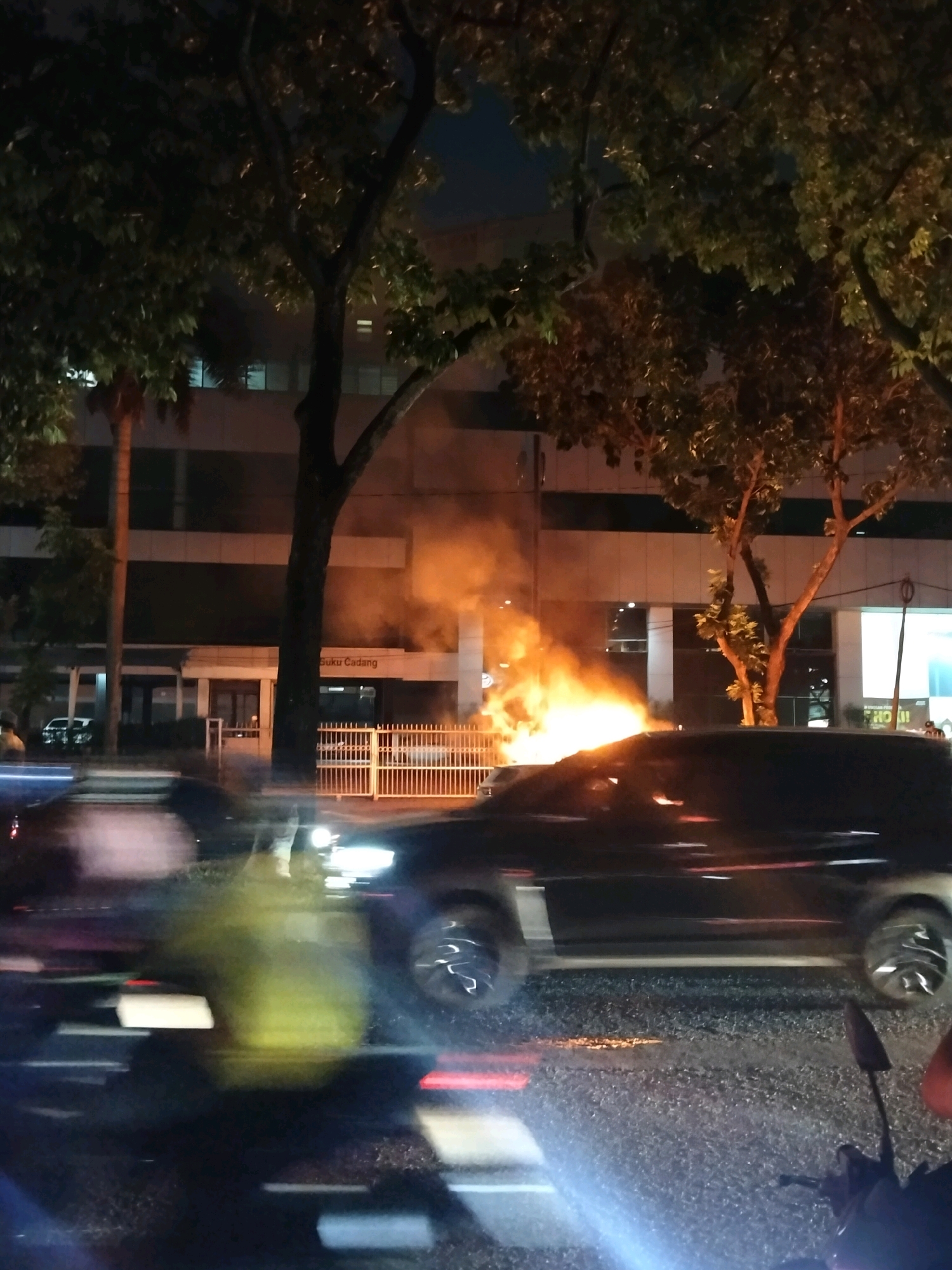 A dark-colored vehicle is engulfed in flames on the side of a road at night in the city of South Tangerang, Indonesia. The fire is producing a large amount of orange smoke and flames that are visible against the background of a building. The building appears to be a commercial establishment, with signage that reads "Suku Cadang". Trees with dark leaves are positioned in the foreground and background, casting shadows. The road is wet, reflecting the lights from the fire and other vehicles. There are other vehicles in motion on the road, creating motion blur. A portion of a motorcycle is visible in the lower right corner of the image. The scene appears to be from a street-level perspective, possibly taken from another moving vehicle.