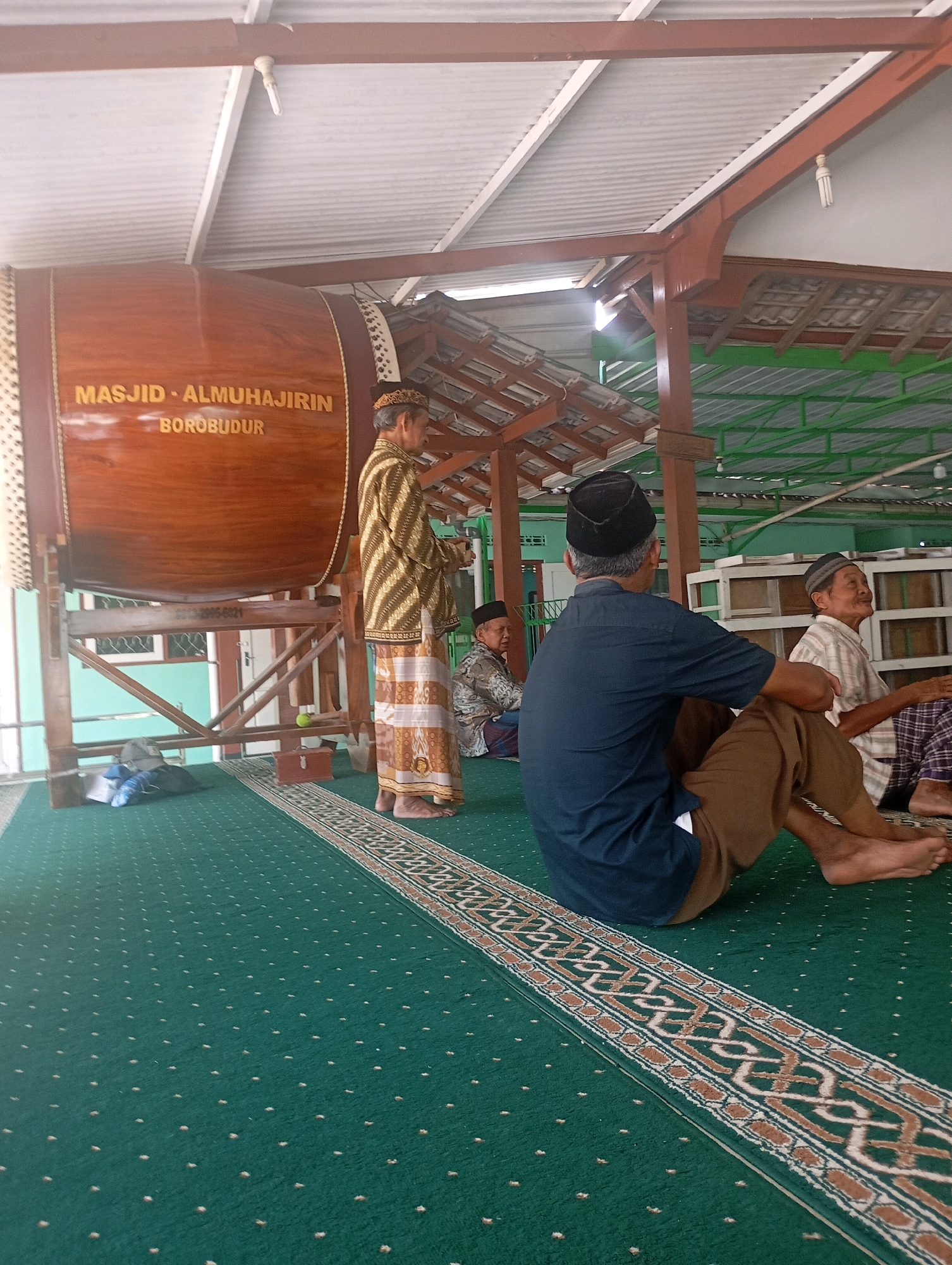The image captures an indoor scene within a building in Borobudur, Indonesia, likely a mosque due to the presence of a large, ornate drum and individuals dressed in traditional attire. The drum, a prominent feature, is made of polished wood and bears Arabic inscriptions identifying it as "MASJID - ALMUHAJIRIN BOROBUDUR." Several men are present; one stands beside the drum wearing a patterned tunic and sarong, while others are seated on a green carpet. One man in the foreground is seen from behind, wearing a dark blue shirt and brown pants, with his legs crossed. Another man sits to his right, wearing a plaid shirt and a skullcap. The environment appears to be an open-air structure with a corrugated metal roof supported by wooden beams. The floor is covered with a green carpet featuring a decorative border. Bags are visible on the floor near the drum. The overall impression is one of a community gathering in a place of worship.