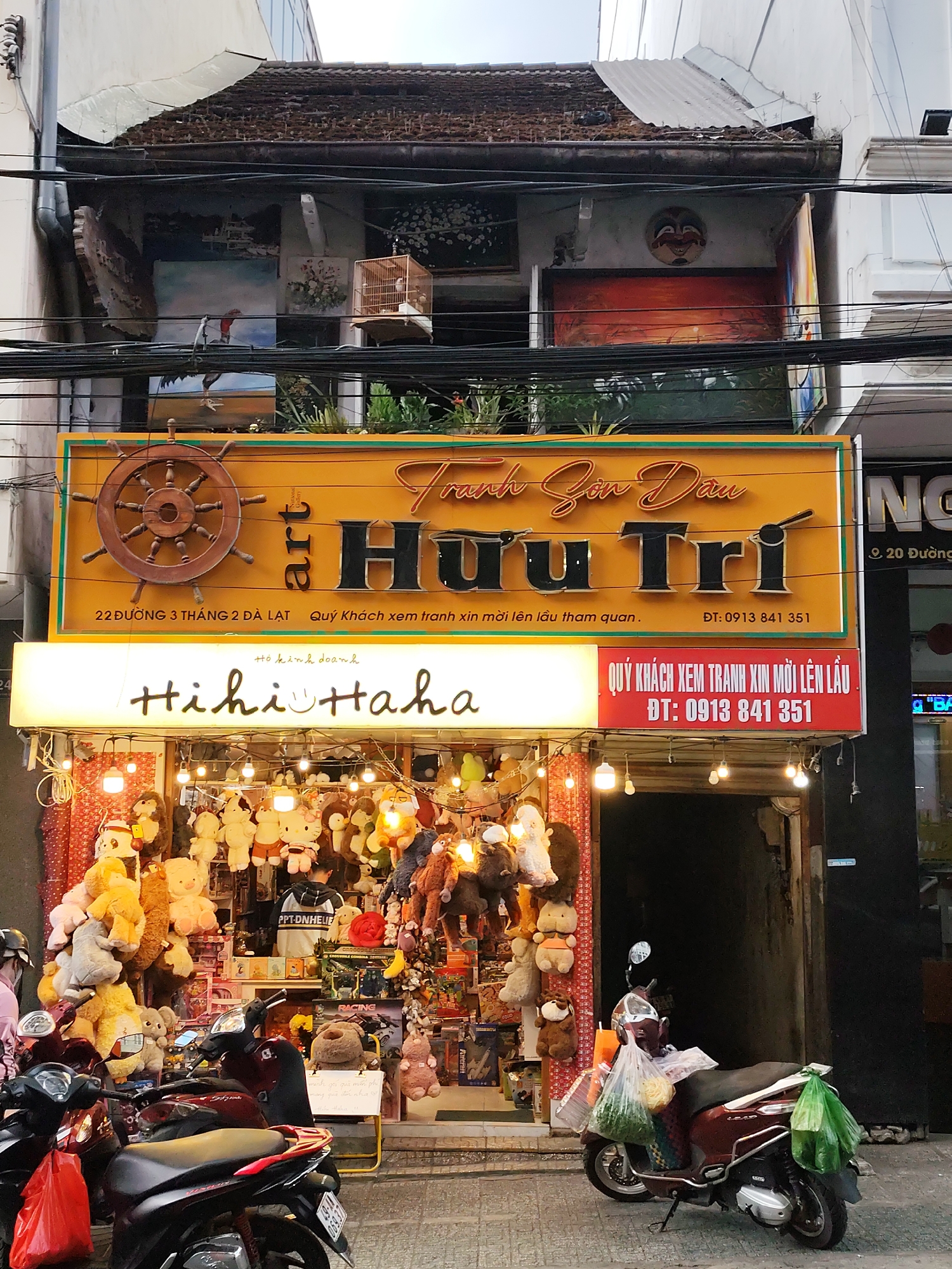 This media file captures a vibrant street scene in Da Lat, Vietnam, featuring a multi-story building housing two distinct businesses. The foreground is dominated by several parked motorbikes, some laden with bags, common in Vietnamese urban settings. Power lines crisscross the upper portion of the image.
The ground floor hosts a brightly lit retail shop named "Hihi Haha," indicated by a glowing sign with a smiley face. Its entrance is densely packed with a colorful array of stuffed animals and plush toys, creating a whimsical and inviting display. Hanging lightbulbs further illuminate the merchandise.
Above this shop, a large yellow sign proudly displays "art Tranh Sơn Dầu Hữu Trí," identifying an oil painting art gallery. The sign also features a wooden ship's wheel and provides the address "22 ĐƯỜNG 3 THÁNG 2 ĐÀ LẠT" and a phone number "ĐT: 0913 841 351." A red sign below reiterates, "QUÝ KHÁCH XEM TRANH XIN MỜI LÊN LẦU" (Dear Customers, please come upstairs to view the paintings) with the same phone number, directing patrons to an upper floor. The building's upper level, with its rustic tiled roof, showcases various paintings displayed on balconies and walls, including a large landscape and a traditional birdcage, hinting at the artistic nature of the establishment. One painting appears to depict a sunset scene, while another shows a boat on water.
The overall impression is one of a bustling commercial street, likely during the day or early evening given the shop's illuminated display and the general ambient light. The combination of the playful toy shop and the more refined art gallery creates a unique juxtaposition in the urban landscape of Da Lat.