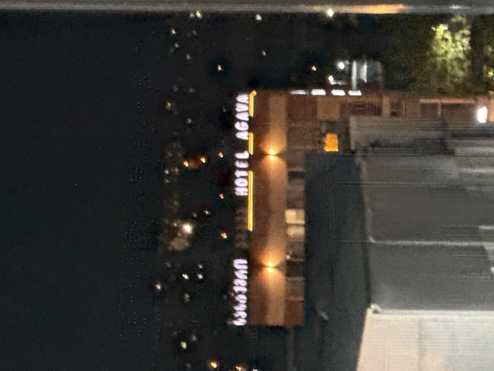 The image captures a nighttime view of the "HOTEL AGAVA" sign in Tbilisi, Georgia. The sign, illuminated with yellow neon lights, is situated above a brick building. To the left of "HOTEL AGAVA" are illuminated characters in Georgian script, though they are not clearly legible. The building has several windows, some of which are lit from within. In the background, a cityscape with numerous scattered lights is visible, suggesting an urban environment. The foreground shows parts of other buildings, including a pale, possibly white, structure on the left and a darker, more textured building on the right. The overall image is slightly blurry due to low light conditions and potentially motion.