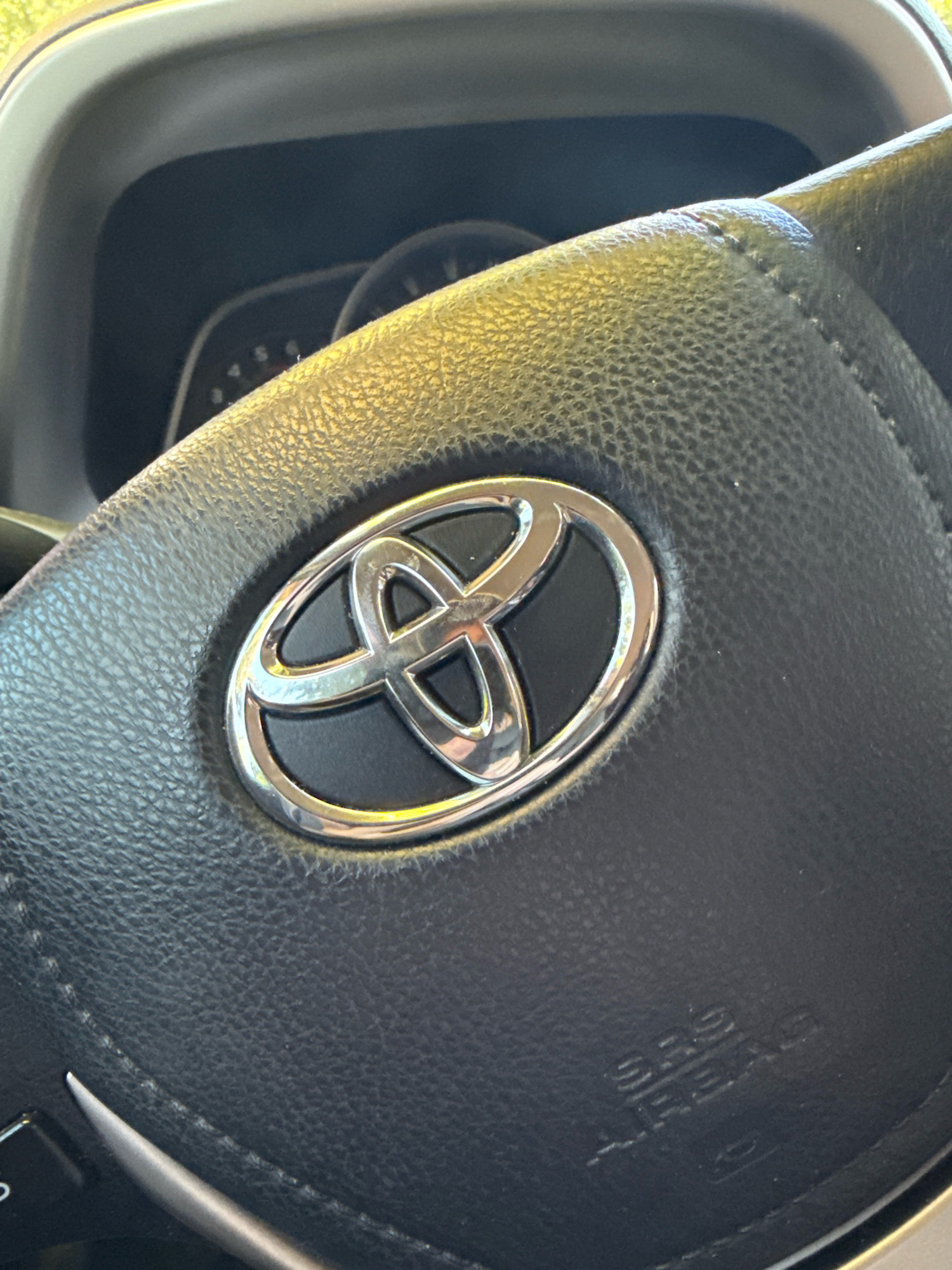 The image is a close-up, eye-level shot of a Toyota steering wheel inside a vehicle. The steering wheel is black and appears to be made of leather. In the center of the steering wheel is the silver Toyota logo, which consists of three overlapping ovals forming a stylized "T". Below the logo, embossed into the leather, are the words "SRS AIRBAG". The dashboard of the car is visible in the background, with some of the instruments slightly blurred. The lighting suggests it is daytime, with natural light illuminating the scene. There are no people or other discernible objects in the immediate vicinity.