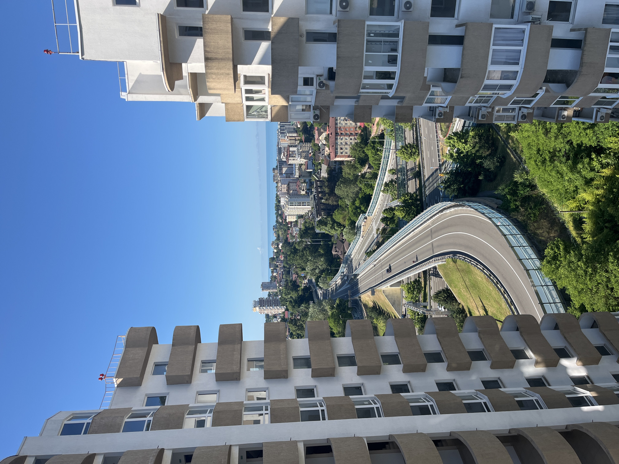 This is an aerial, eye-level, long-shot photograph taken on a clear, sunny day in Sochi, Russia. The image is dominated by two modern, multi-story apartment buildings on the left and right edges, with a highway curving through the center of the frame. The buildings are white with protruding balconies, some of which have curved, beige accents. The balconies are uniformly spaced and appear to have large windows. Air conditioning units are visible mounted on the exterior walls of the buildings.
The highway is a multi-lane road that curves from the foreground into the distance, elevated in parts and surrounded by green grassy slopes and trees. A few vehicles are visible on the highway. In the background, a dense urban landscape stretches towards the horizon, characterized by a mix of mid-rise and high-rise buildings, interspersed with lush green trees. Beyond the city, the blue expanse of the sea is visible, meeting the clear blue sky at the horizon. Two small red objects, possibly flags or markers, are visible on the rooftop of the building on the right. The overall scene depicts a modern urban environment in a coastal city.