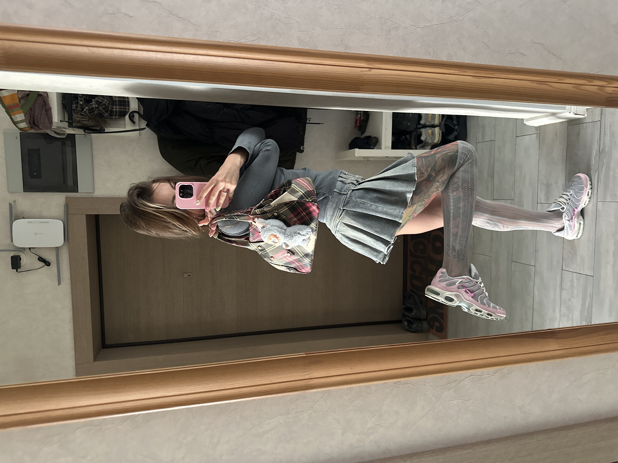 A woman stands in what appears to be a hallway, taking a selfie in a mirror. She is wearing a grey long-sleeved shirt, a grey pleated mini-skirt, white patterned stockings, and pink and grey sneakers. She is carrying a plaid purse with a stuffed animal keychain. Tattoos are visible on her legs. In the background, a door, shelves with clothing and shoes, and a router are visible. The floor is tiled. The image appears to have been taken in Moscow, Russia, based on the user's prompt.