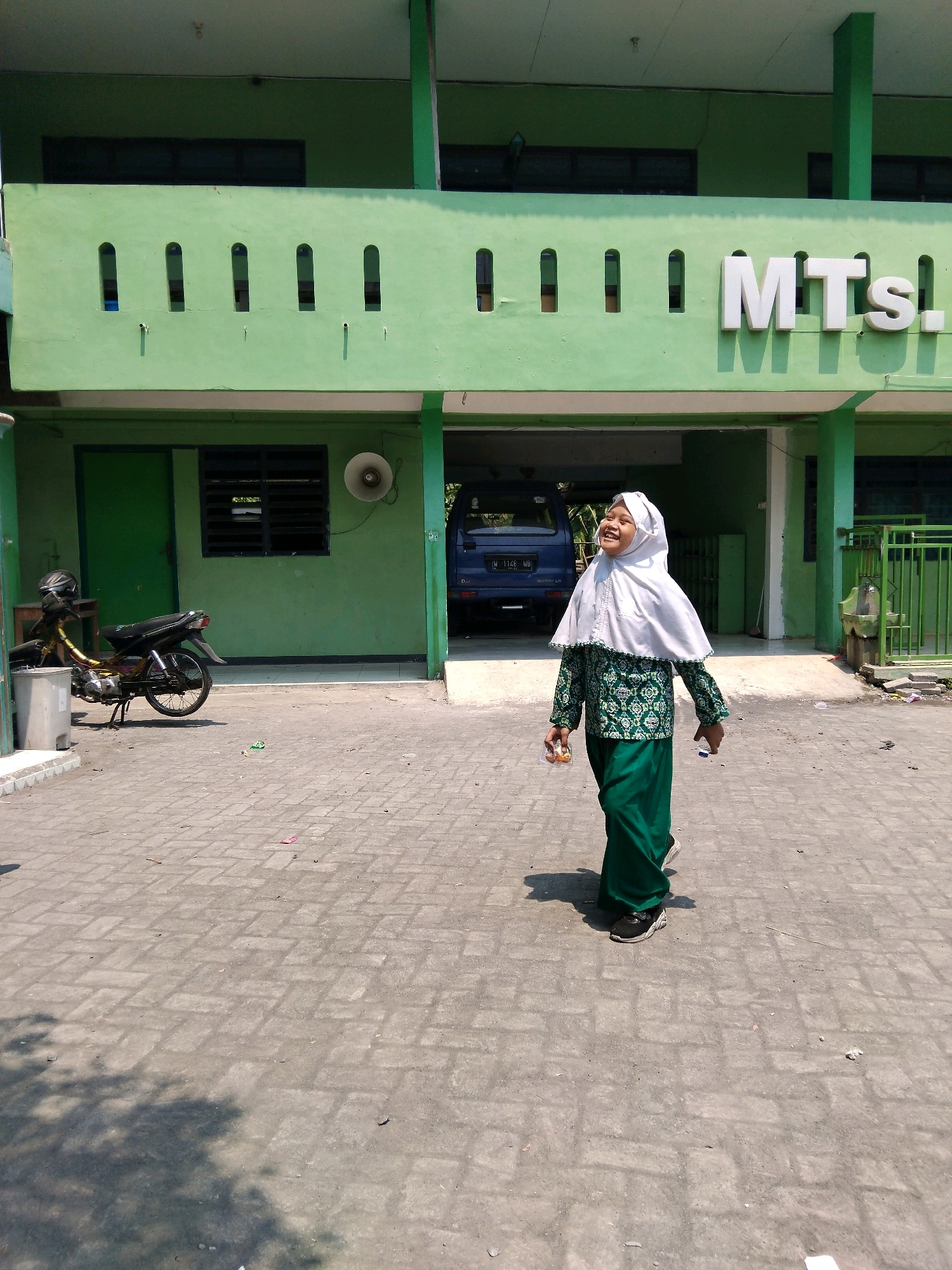 A young girl wearing a white hijab and a green patterned dress stands on a paved courtyard in front of a light green building in Kepuh Kiriman, Indonesia. The building has the letters "MTS." in white lettering above an arched opening. A blue van is parked in the opening. A motorcycle is parked to the left of the opening, and a trash can is beside it. The girl is smiling and looking upwards. She is holding a small plastic package in her right hand. The ground is paved with interlocking concrete blocks. Sunlight casts shadows of trees on the ground.