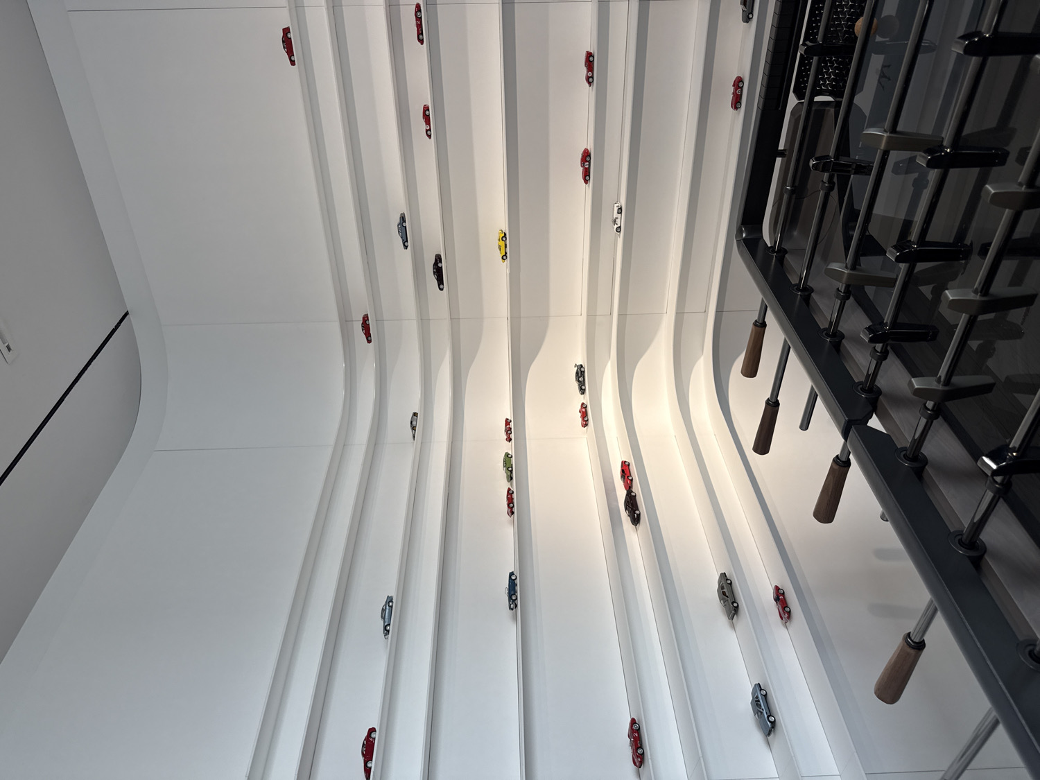 This high-angle image captures a striking modern indoor display, likely in Milan, Italy, featuring numerous miniature model cars arranged on flowing, white architectural elements that resemble race tracks. The perspective looks sharply downwards, suggesting these elements might be on a high wall or even the ceiling.
The main subjects are the dozens of small, detailed model cars of various colors, including red, yellow, green, blue, black, grey, and white. They are meticulously placed along long, parallel, undulating white tracks that are integrated into the predominantly white surface of the room. The wavy design of these tracks creates a dynamic sense of motion, as if the cars are in a continuous race or procession.
To the right of the frame, a significant portion of a modern foosball table is visible. Its dark grey or black structure, shiny player rods, and distinct wooden handles are clearly depicted, adding a contrasting element of recreation to the sleek display. The playing surface of the foosball table is dark, reflecting some light.
The setting appears to be a contemporary and minimalist interior space, possibly a showroom, museum, or office, characteristic of Milan's design aesthetic. The lighting is bright and even, indicating artificial indoor illumination, and there are no visible external light sources or weather conditions. No people are present, so there is no direct activity taking place, but the arrangement implies a permanent exhibit or artistic installation. The foosball table suggests a leisure or interactive zone. No visible text is present in the image. The overall impression is one of clean lines, modern design, and playful sophistication.