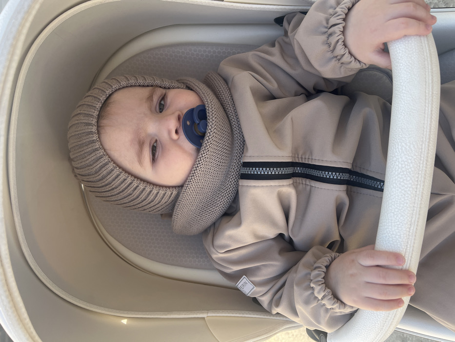 An infant is seated in a light beige or off-white stroller in the city of Noviy, Russia. The infant, approximately 6-18 months of age, wears a light brown ribbed knitted hat, a matching knitted snood, and a light brown jumpsuit with a central black zipper and reflective grey stripes. A small "PERINI" label is visible on the jumpsuit's left chest. A dark blue pacifier is held in the infant's mouth. The infant's right hand rests on the stroller's white padded bumper bar. The background is brightly lit and out of focus, indicating an outdoor environment.