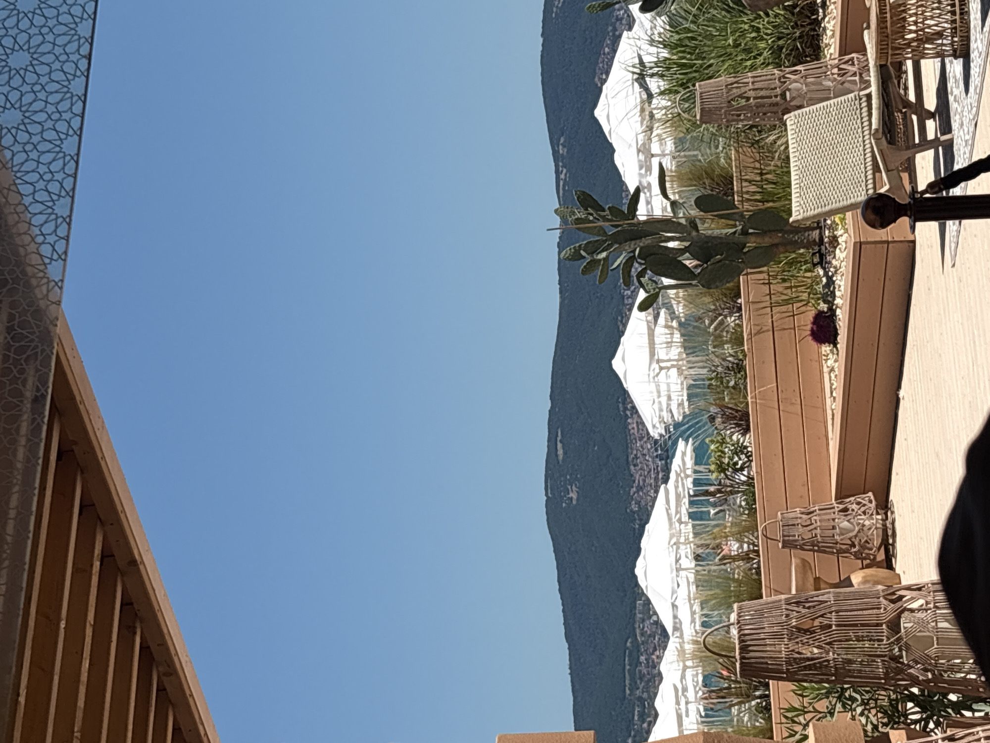 The image shows a resort area on a sunny day with a clear blue sky. In the foreground, there are two large wicker lanterns and a woven chair. Behind them, a wooden deck with planters filled with greenery and a cactus is visible. In the middle ground, rows of white beach umbrellas are set up on the sand, with a glimpse of the sea and a town with red-tiled roofs nestled at the foot of a large, tree-covered hill in the background. The location is identified as Nesebar, Bulgaria.