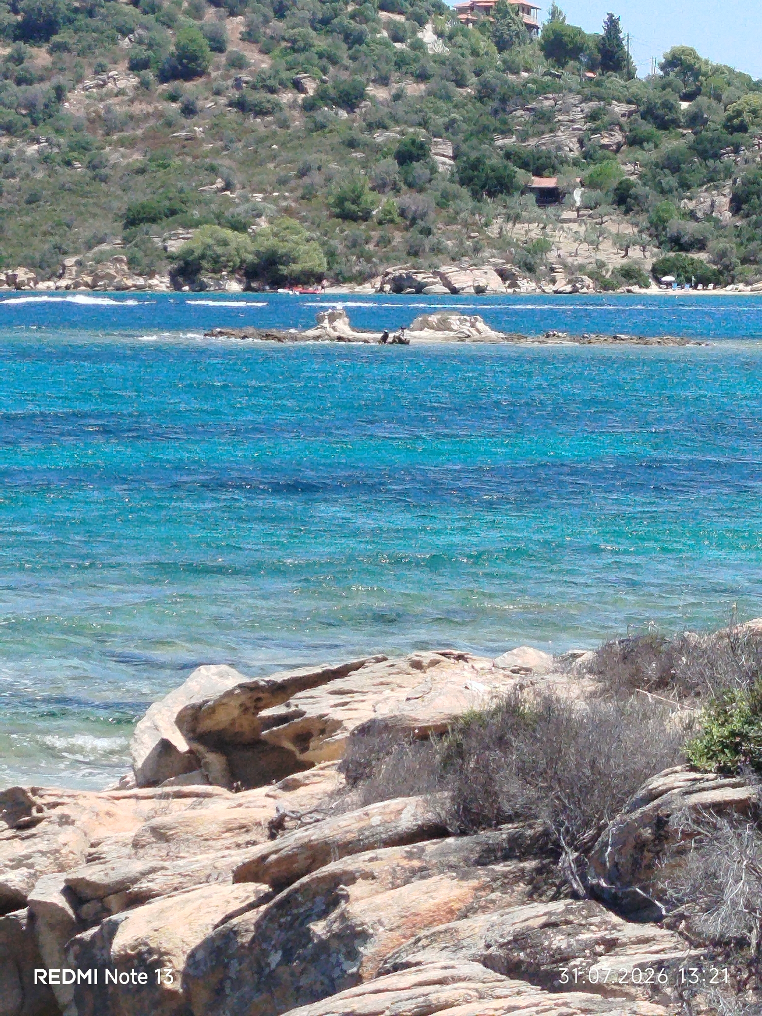 This is a photograph of a scenic coastal view in the Sithonia Municipal Unit, Greece. The image captures a vibrant blue sea with clear turquoise waters near the shore. Rocky formations are present in the foreground, with some sparse vegetation. In the mid-ground, a small rocky island with a few people on it is visible. The background features a lush, green hillside dotted with trees and some buildings, suggesting a residential or resort area. The sky is a clear, pale blue. A timestamp and the text "REDMI Note 13" are present in the lower left and lower right corners of the image, respectively.