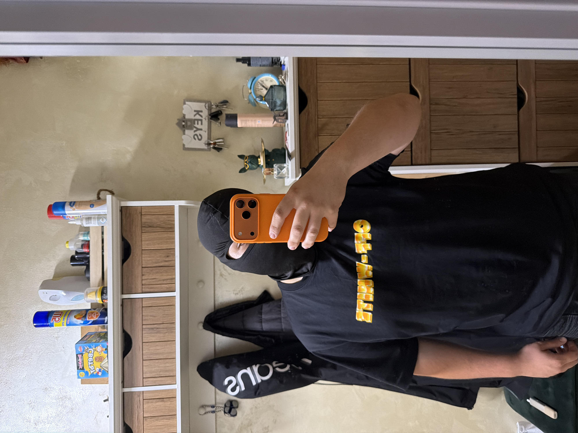 This mirror selfie, taken indoors in what appears to be an entryway or hallway in Sofia, Bulgaria, features an individual wearing a black balaclava covering their face, with only the eyes visible. The person is dressed in a black short-sleeved t-shirt, prominently featuring the reversed text "Off-White™" in a yellow and orange gradient across the chest. They are holding an orange smartphone, likely an iPhone due to its distinctive camera module, in their right hand to capture the reflection. A black jacket or hoodie is visible near the person's left arm, with the reversed word "snaps" faintly discernible on an attached tag or bag.
The background reveals a light-colored, possibly textured, wall. On the wall, a white key holder shaped like a house displays the reversed word "ƧYƎ⋊" (KEYS) and holds several keys. Beneath it, a white and wooden shelving unit houses various items, including a light blue alarm clock, several bottles or containers, and a green decorative cat figurine wearing a hat. To the left, another white shelving unit with wooden drawers contains household products like a blue spray bottle with partially visible text "MR D...", a white spray dispenser, and a board game box with "CBBG..." printed on it. The lighting suggests an indoor setting, with no specific time of day indicated. No emotions are discernible due to the balaclava.