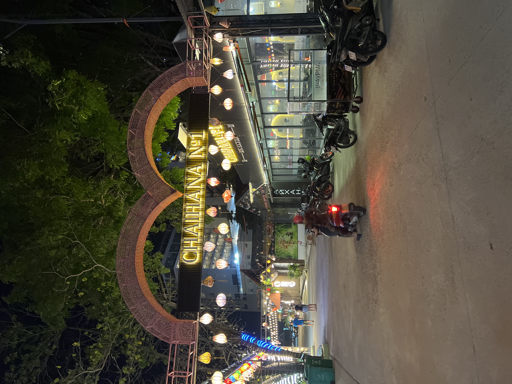 The image depicts a nighttime street scene in Nha Trang, Vietnam, featuring a prominent archway decorated with lights and lanterns. Underneath the arch, a neon sign reads "CHAIHANA Nº1". The street is paved and lined with buildings, some of which appear to be businesses with large glass windows. Several motorcycles are parked on the right side of the street, and one person is riding a red motorcycle in the foreground. In the distance, a few pedestrians are walking. Trees with lush green foliage are visible above the archway, and decorative lanterns are suspended from wires, illuminating the street. The overall atmosphere is festive and vibrant, typical of a commercial area at night.