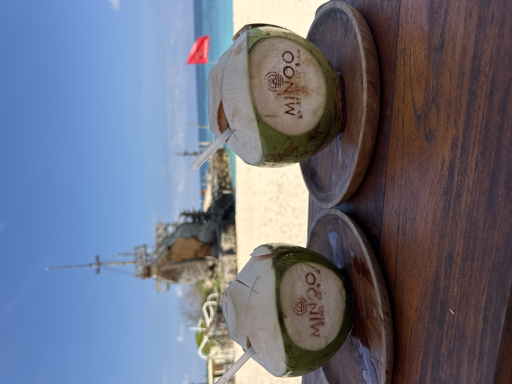 Two fresh, green coconuts, each with a white straw, are positioned in the foreground on a dark wooden table. They rest on individual round wooden coasters, with some liquid visible on the coasters, suggesting condensation or spillage. The coconuts are branded with a logo featuring a wave-like symbol and the text "MINOO BEACH CLUB."
Behind the table, a bright, sandy beach stretches towards the clear blue ocean. A red flag is visible flying near the water's edge on the left side of the frame. Further back and slightly to the left, a large, dark-colored wooden structure resembling a ship or boat (possibly a pirate ship replica) with prominent masts and rigging is partially visible under a clear blue sky.
The scene depicts a sunny daytime setting, suggesting a leisurely vacation or relaxing activity at a beach club. The overall atmosphere is tropical and bright. The location is Kuta Selatan, Indonesia.