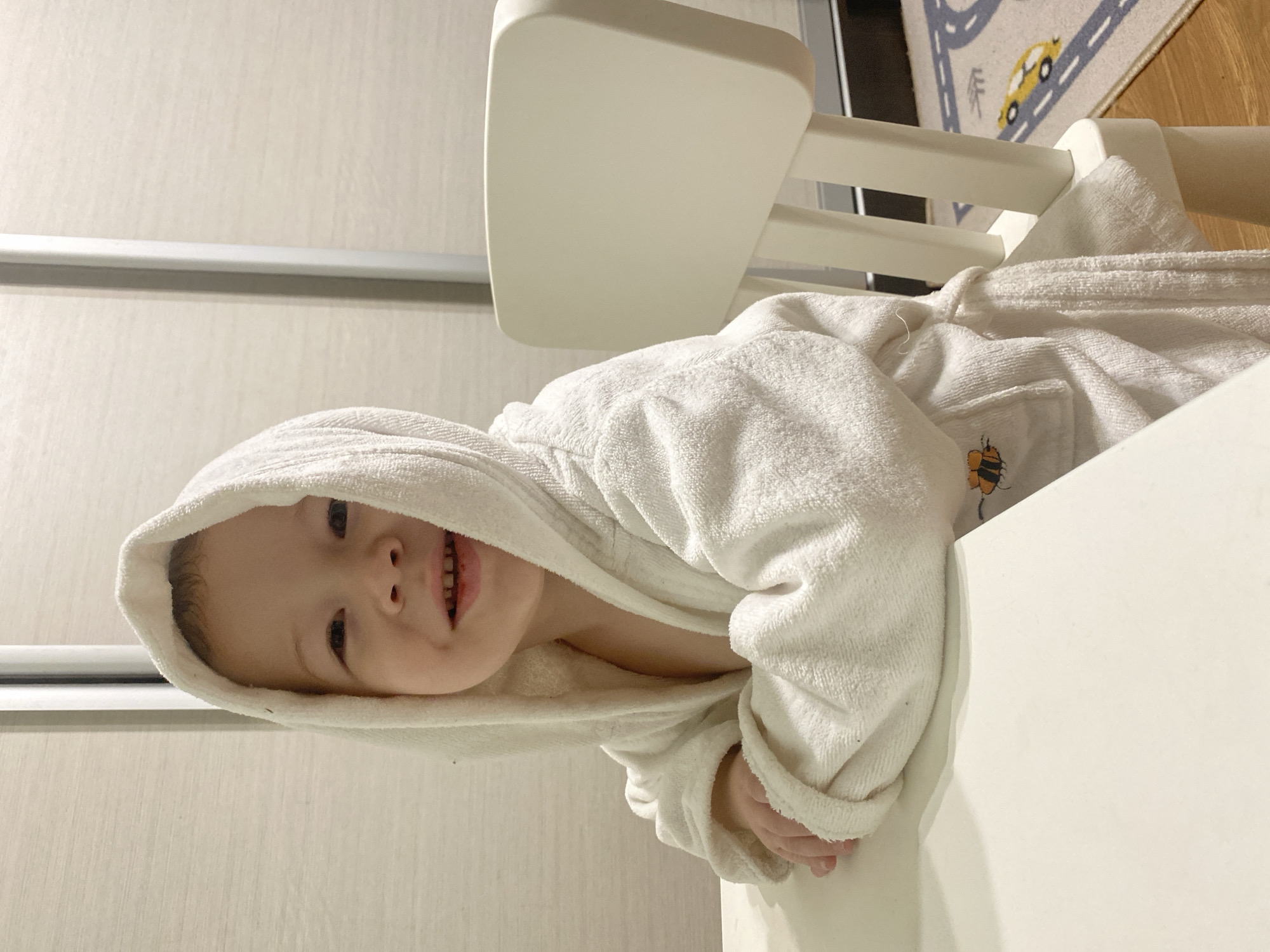 A child wearing a white hooded bathrobe sits in a white chair, smiling. A portion of a rug with a car design is visible in the background. The location cannot be determined from the image.