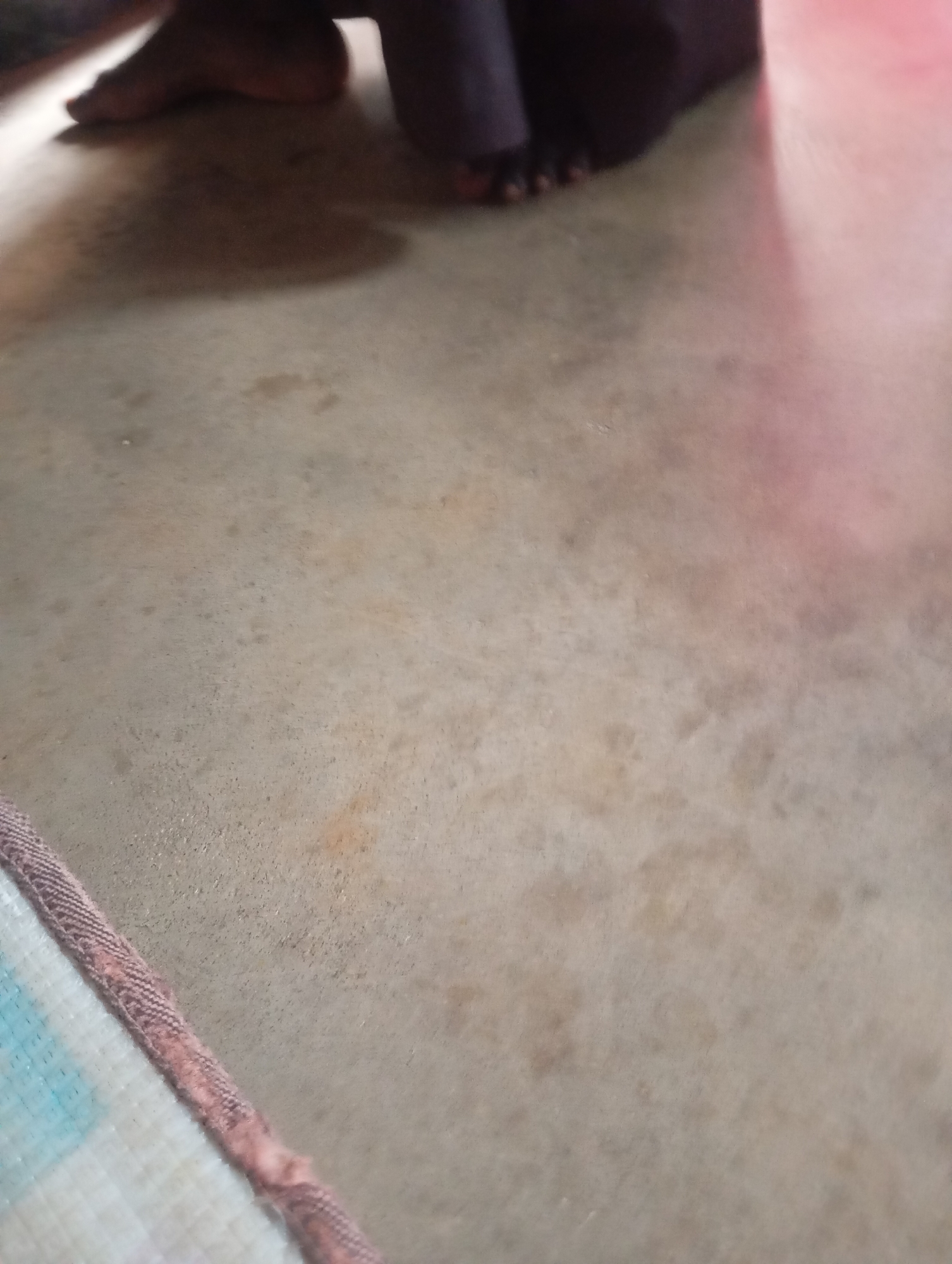 The image shows a close-up view of a person's bare feet and lower legs standing on a concrete floor. The feet are dark-skinned and appear to be positioned slightly apart. The concrete floor is a light grey color with some darker patches and stains, suggesting it is an outdoor or utilitarian space. In the lower left corner, a portion of a woven mat or rug with a patterned edge is visible. The lighting suggests it might be daytime, with some shadows cast by the person's legs. The overall scene is simple and focuses on the immediate ground level. There are no discernible landmarks or urban features to indicate a specific location within Jalingo, Nigeria, beyond the context provided.