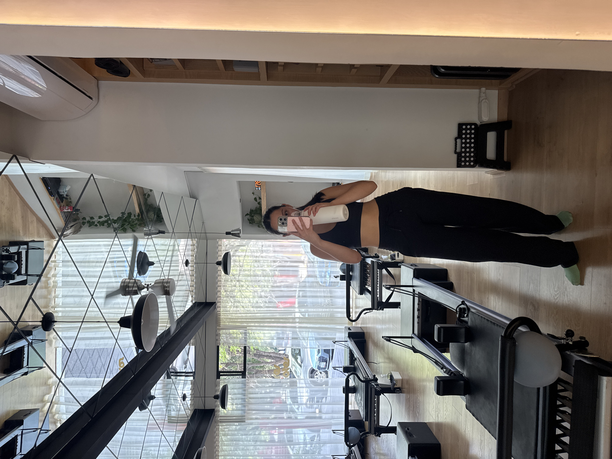 A woman wearing a black sports bra and black pants stands in what appears to be a pilates studio, holding a white water bottle and taking a selfie with a pink phone. The pilates studio contains several pilates reformers arranged in parallel. Light streams in through large windows covered with sheer white curtains. Outside the windows, trees and cars are visible. The studio has wooden floors and a high ceiling with modern, geometric black light fixtures. A white air conditioning unit is installed near the ceiling. A wooden bookshelf and a small black folding step stool are also visible near the ceiling. This could be a pilates studio located in Puebla, Mexico, based on the style of architecture and decor.