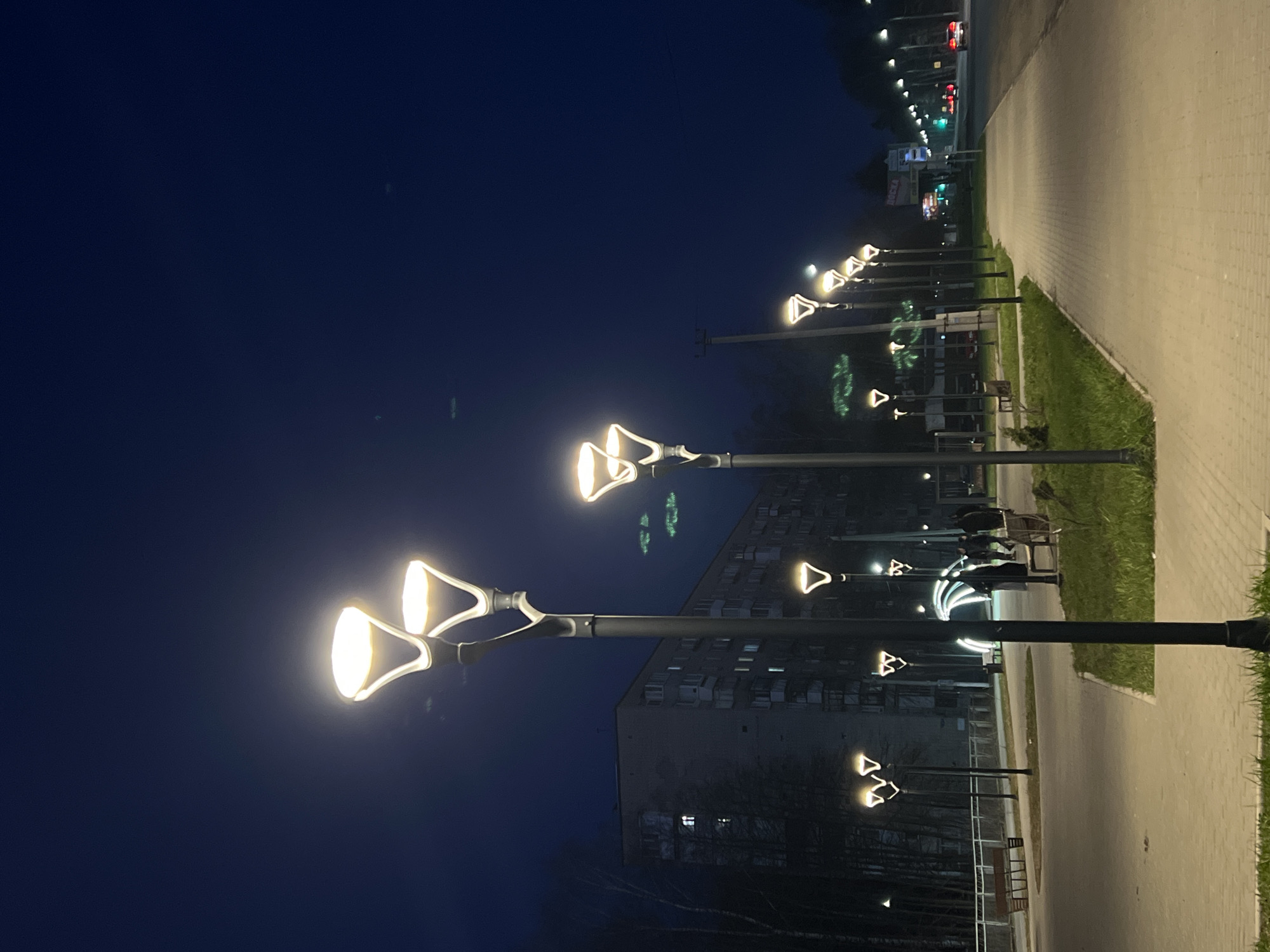The image displays a nighttime urban scene, likely a pedestrian area alongside a street in Chaikovsky, Russia. The perspective is oriented to show a paved pathway on the right, made of light-colored blocks, running parallel to a grassy verge. To the left of the grass strip is a darker, possibly unpaved road or another pathway. Further in the background, a large, multi-story building, likely an apartment complex, is visible with some illuminated windows. Bare trees are present between the building and the foreground elements, suggesting a late autumn or winter setting.
The main subjects are a series of modern streetlights with distinctive designs. Each streetlight features two bright, warm-white luminaires that are triangular or flared-cone shaped, casting a strong glow on the immediate surroundings. These lights extend in a line into the distance, illuminating the path and green spaces. In the far distance, along a main road, other vehicle lights (red taillights and possibly headlights) are faintly visible, indicating traffic. Several benches can be seen in the grassy areas, suggesting a public park or walkway. The sky is a deep, clear dark blue, indicating the late hour. No people are visible, and no specific activity is taking place; the scene appears quiet and still. Faint, blurry Cyrillic text is discernable on a distant sign, but it is illegible.