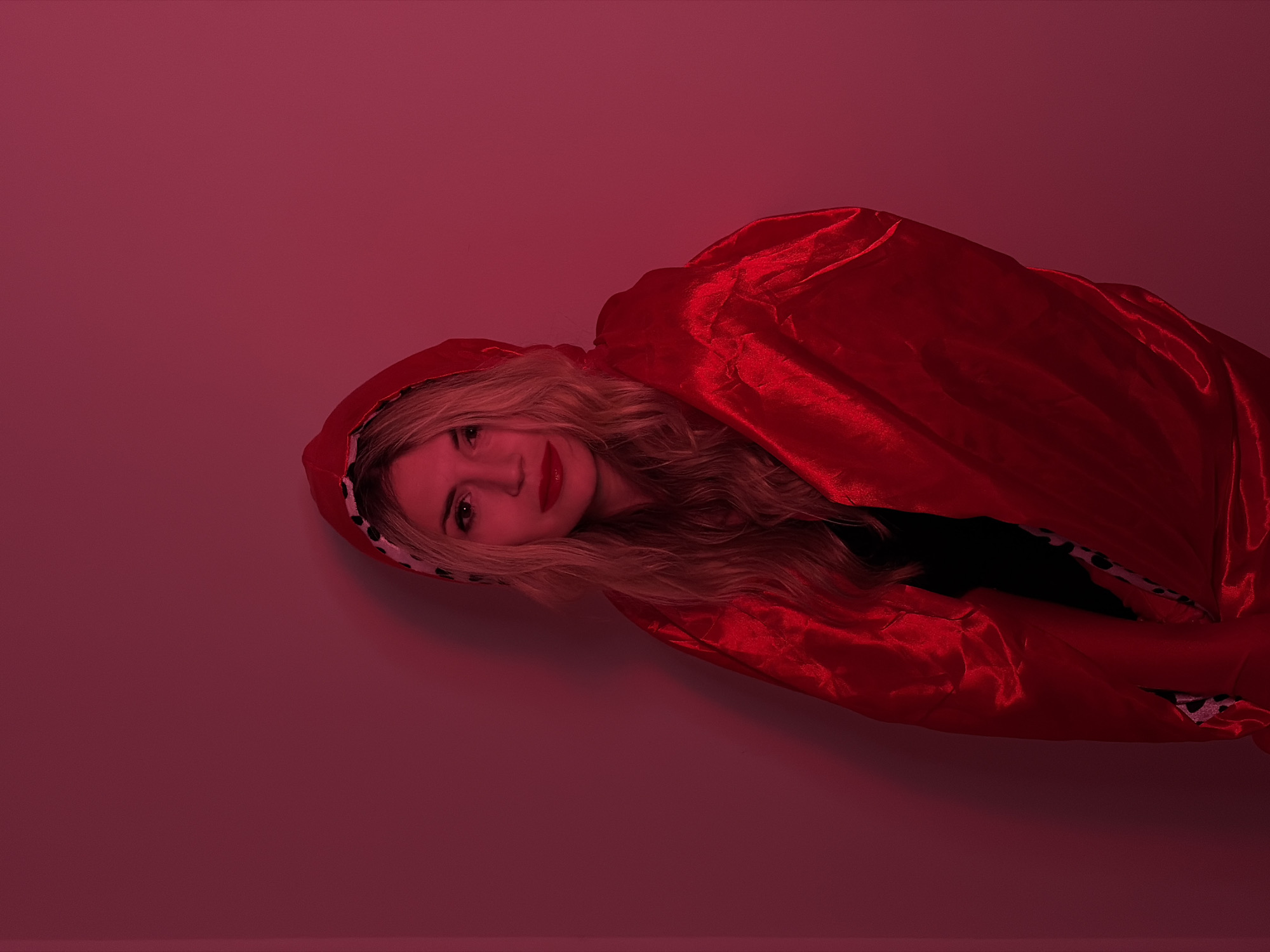 A woman with blond wavy hair is lying down in a hooded red cloak. Her head is slightly tilted and she is looking at the camera. The background is a uniform red color. There are no discernible elements to suggest the location is Budva, Montenegro.