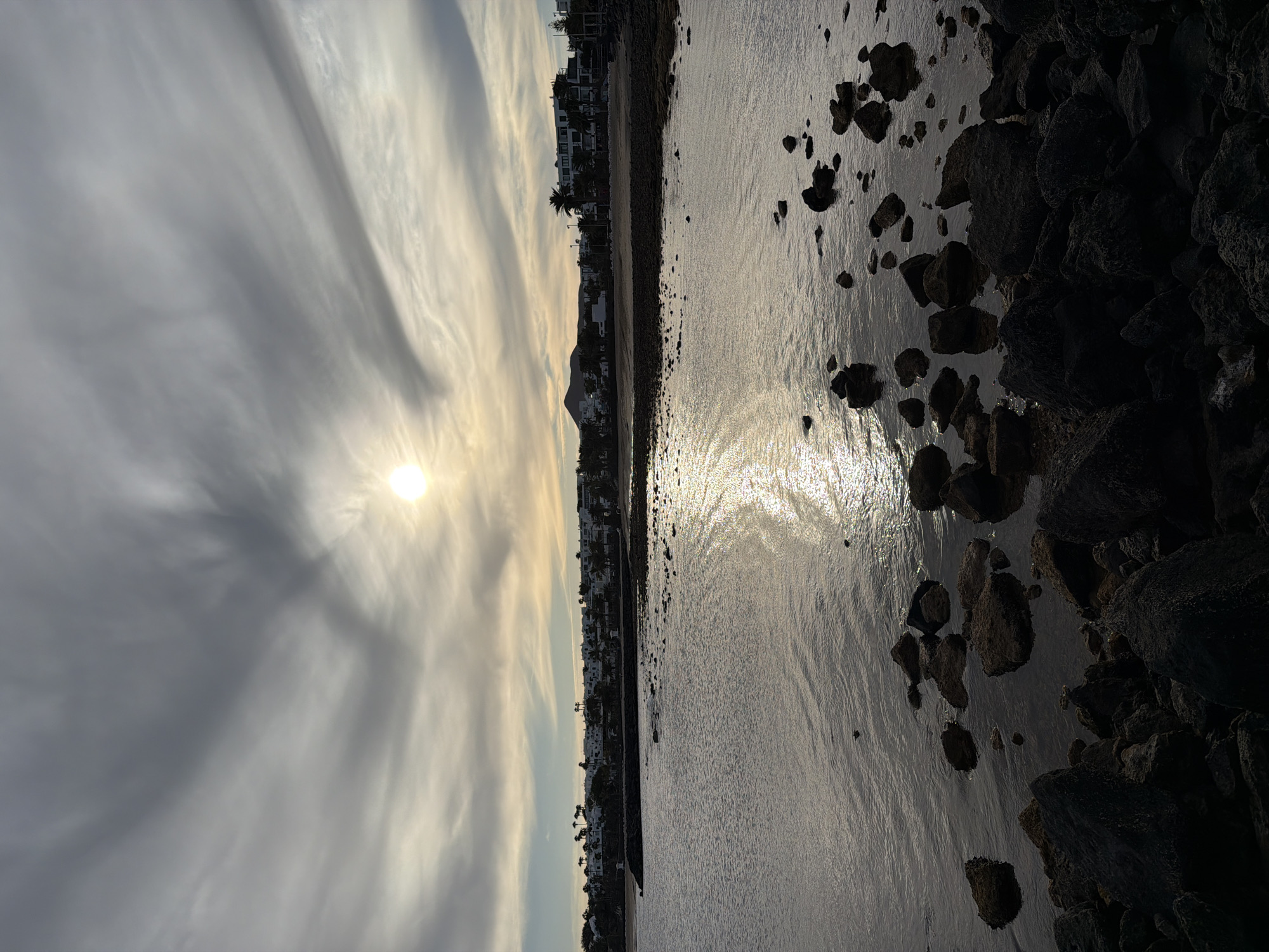 This image captures a serene coastal scene at what appears to be late afternoon or early morning, given the low angle and bright reflection of the sun. The setting is likely a beach or rocky shoreline in Teguise, Spain, consistent with the characteristic white buildings and volcanic rock formations of Lanzarote.
The sky occupies the upper portion, featuring wispy, elongated clouds that stretch across the frame, partially obscuring the bright, circular sun. The sun's light pierces through the clouds, casting a brilliant glow that extends into a shimmering path across the water.
Horizontally bisecting the image is the coastline. A row of white buildings, possibly houses or hotels, stands along the shore, interspersed with tall palm trees. In the distance, a small mountain or hill is visible, silhouetted against the lighter sky.
The foreground is dominated by the calm waters, which reflect the sun's sparkle. Scattered dark, rugged rocks of varying sizes are visible both on the wet sand at the edge of the water and partially submerged. These rocks create textural contrast with the smooth water surface. No people or specific activities are visible, emphasizing the natural landscape and the play of light. The overall mood is tranquil, with muted tones of grey, silver, and soft gold from the sunlight. There is no visible text in the image.
