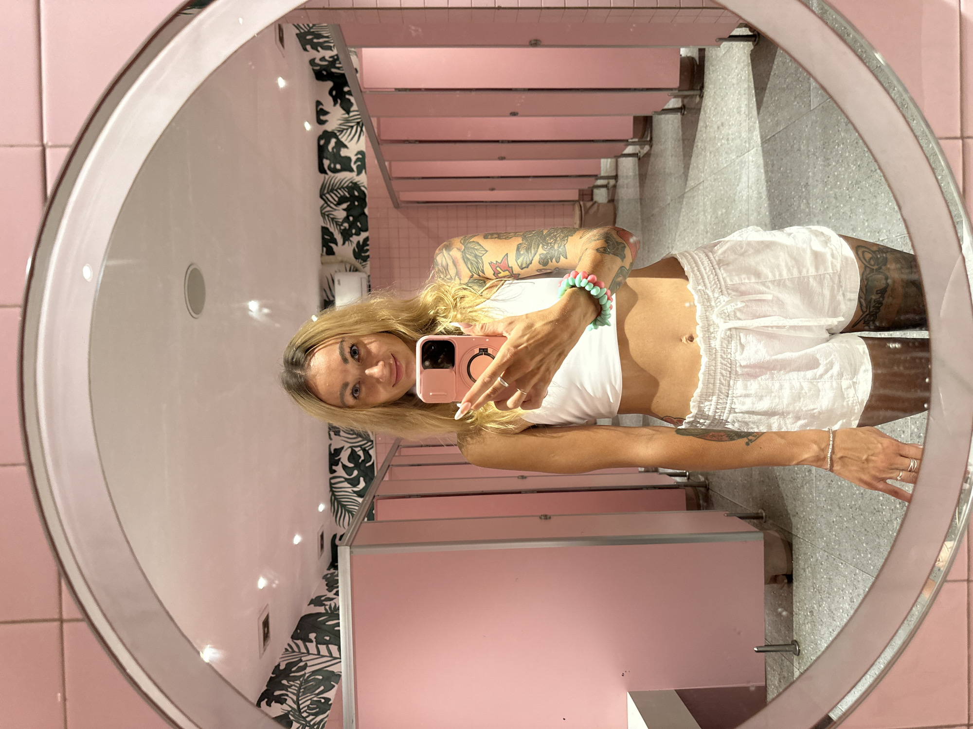 A woman is captured in a mirror selfie, with the image rotated 90 degrees clockwise. She has long blonde hair and is smiling towards her phone. She is wearing a white crop top and white shorts, with extensive tattoos visible on both arms and legs. On her left wrist, she wears a bracelet with pink and turquoise beads, and on her right wrist, a silver chain bracelet. She also has multiple rings on her fingers and long, light-colored fingernails. She holds a light pink smartphone with a circular pop socket or holder on the back.
The setting appears to be a public restroom, likely located in Tien Thanh Ward, Vietnam. The reflection in the large, round mirror shows walls tiled in a soft pink color. There are several bathroom stalls with matching pink doors and partitions, and the floor is a light gray, speckled material. Portions of the wall feature a decorative pattern of large, dark green tropical leaves, resembling monstera. The space is well-lit with indoor artificial lighting. No text is visible in the image.