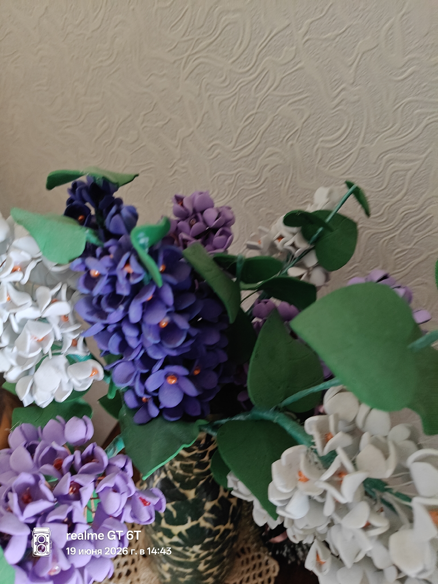 A bouquet of artificial flowers, including purple lilacs and white flowers, is displayed against a textured, light-colored wall. Green leaves are interspersed among the blooms. A small camera icon and text reading "realme GT 6T 19 июня 2026 г. в 14:43" are superimposed on the lower left corner of the image. The image does not provide any information about the location in Tbilisi, Georgia.
