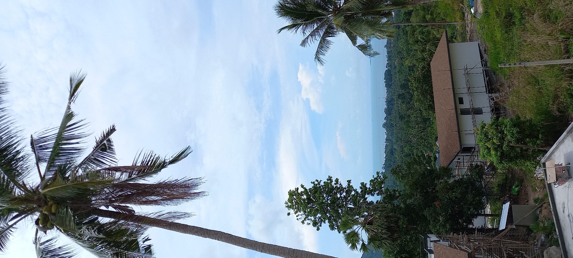 Baan Tai, Thailand: A partially obscured view of the sea and lush tropical forest is framed by coconut palm trees. A two-story building under construction with scaffolding is visible on the right side of the frame. The sky is light blue with scattered clouds.